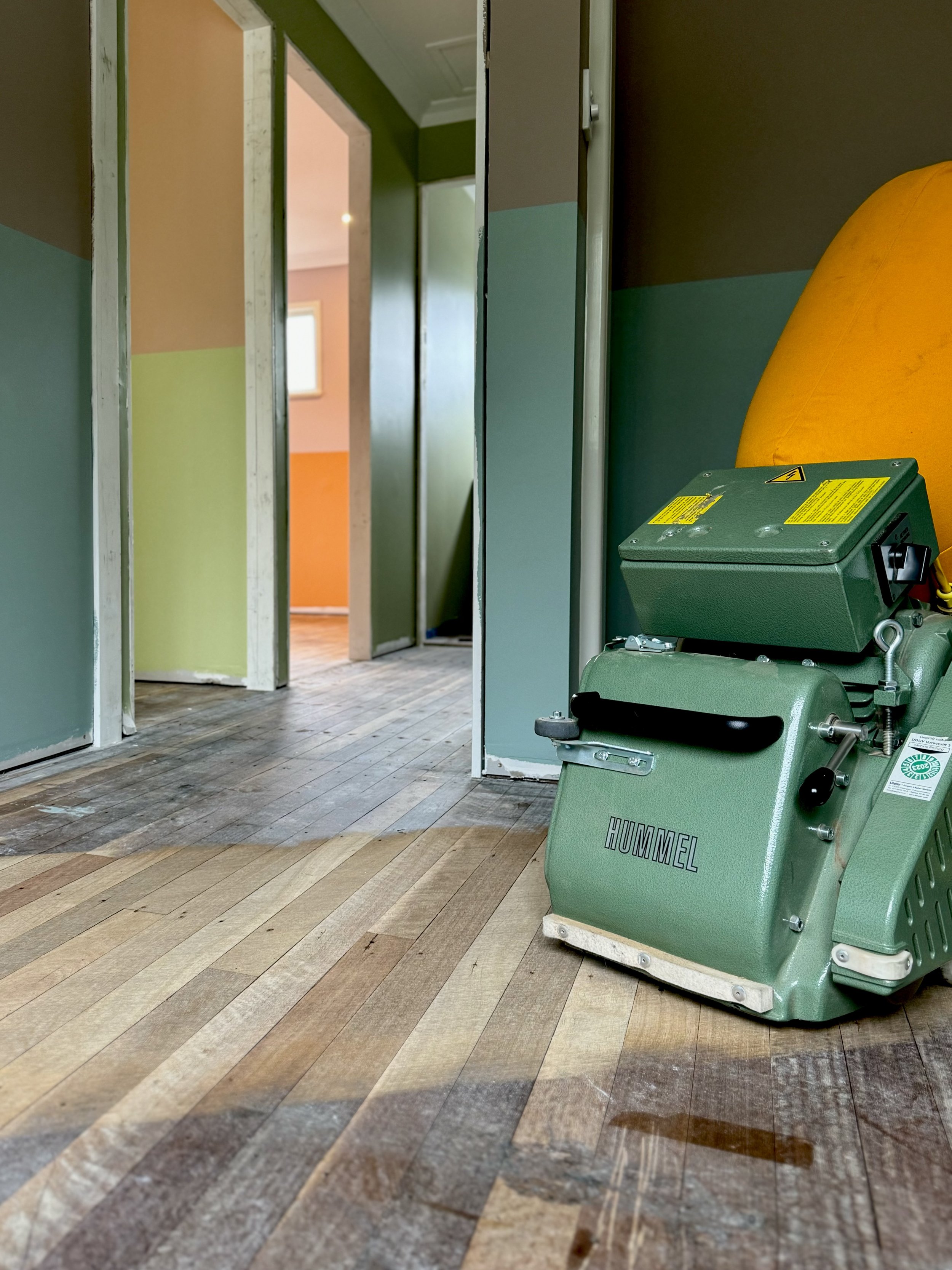 A green commercial floor sander on a wooden floor in a building under renovation.