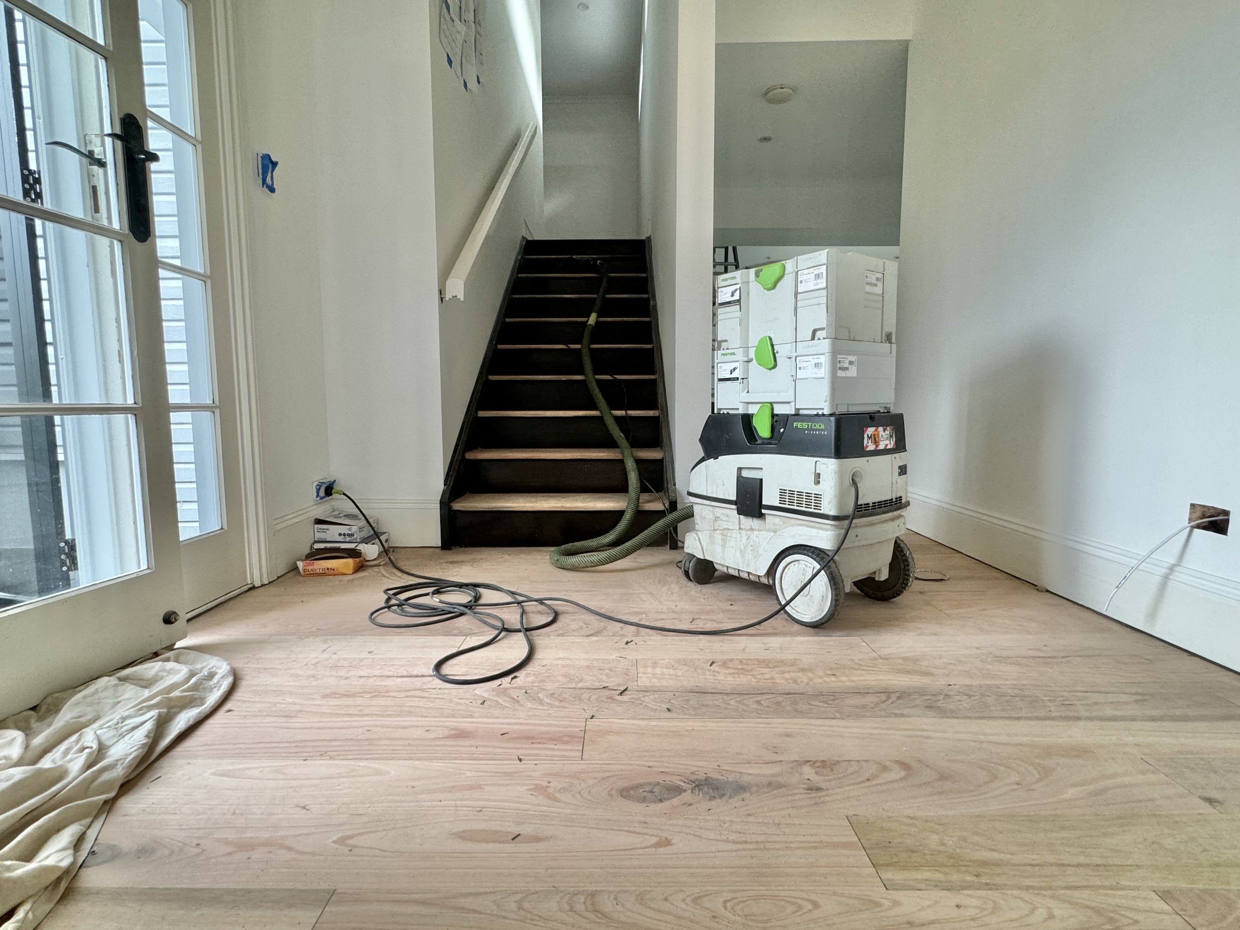 Empty room with hardwood floor and unfinished electrical outlets, containing construction equipment, supplies, and a staircase leading to an upper level.