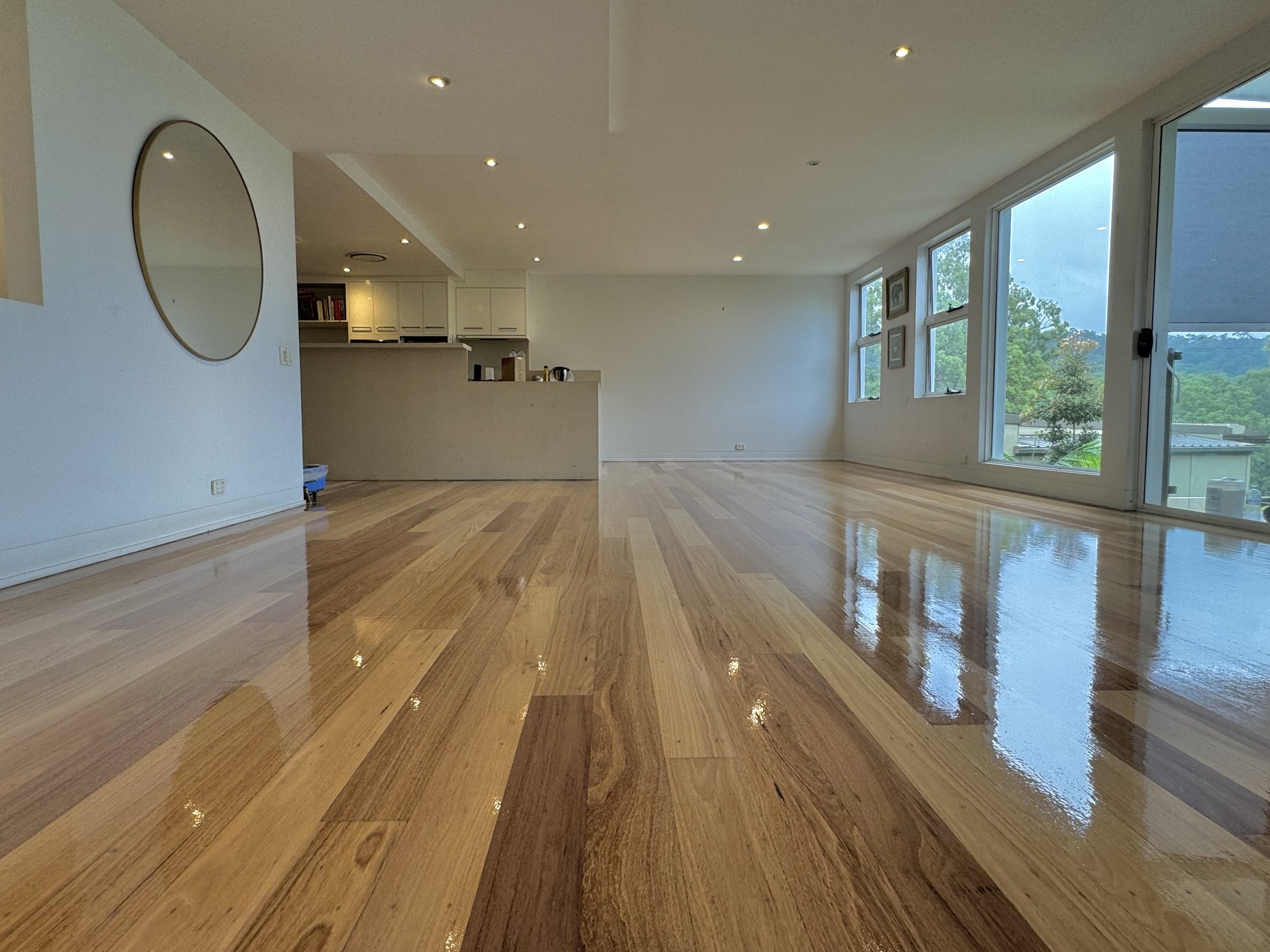 Empty living room with polished wooden floors, white walls, large windows with scenic view, and a kitchen area in the background.
