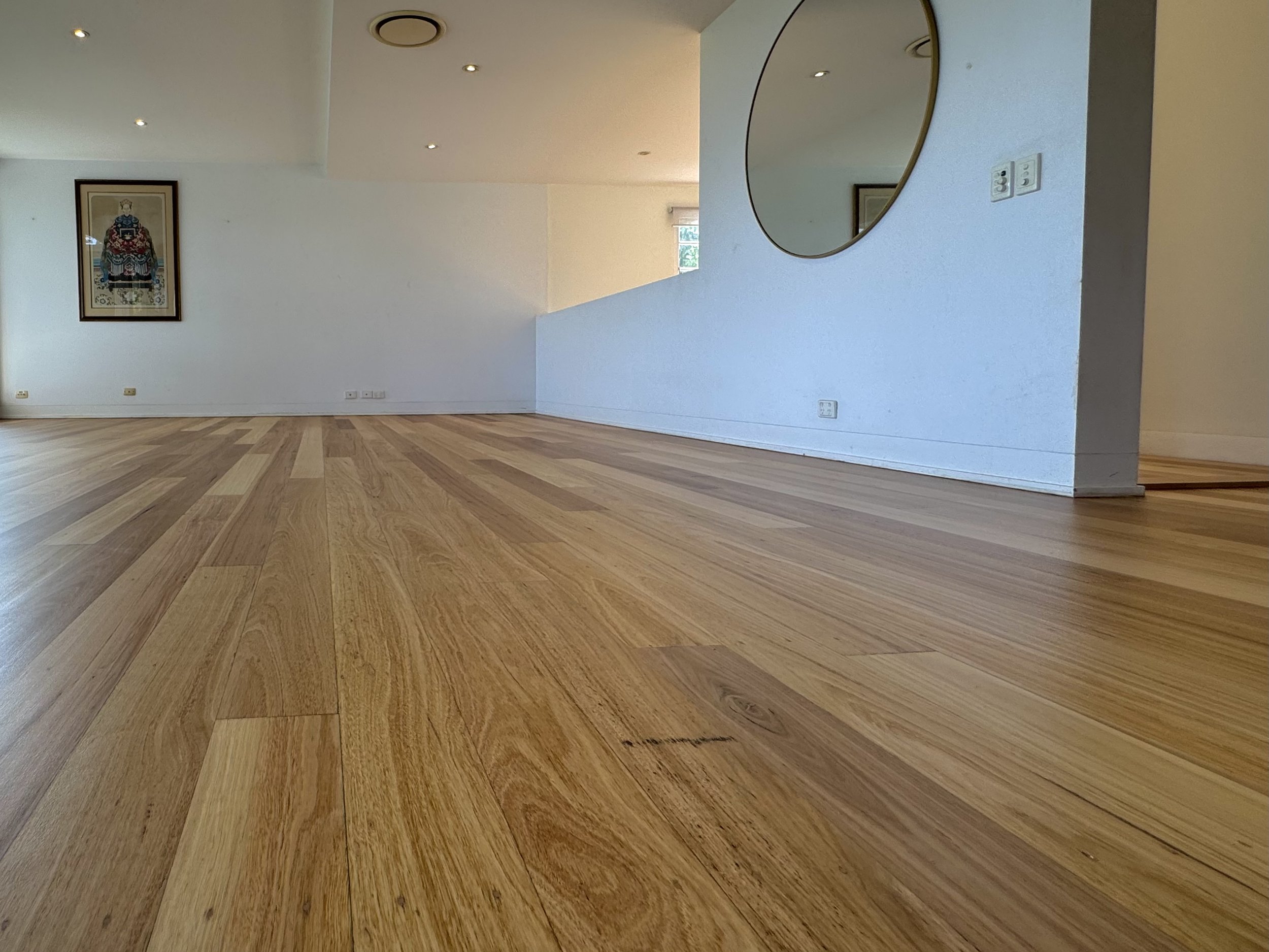 Empty room with wooden flooring, a white wall with a large round mirror and a framed artwork, and a window letting in natural light.