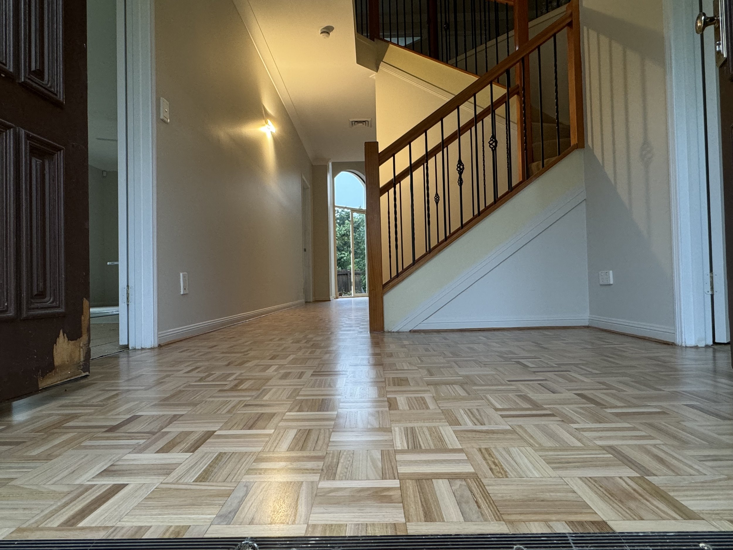 Empty living space with wooden floors, a staircase with wooden railing and black metal spindles, small wall-mounted lights, and window at the end of hallway.