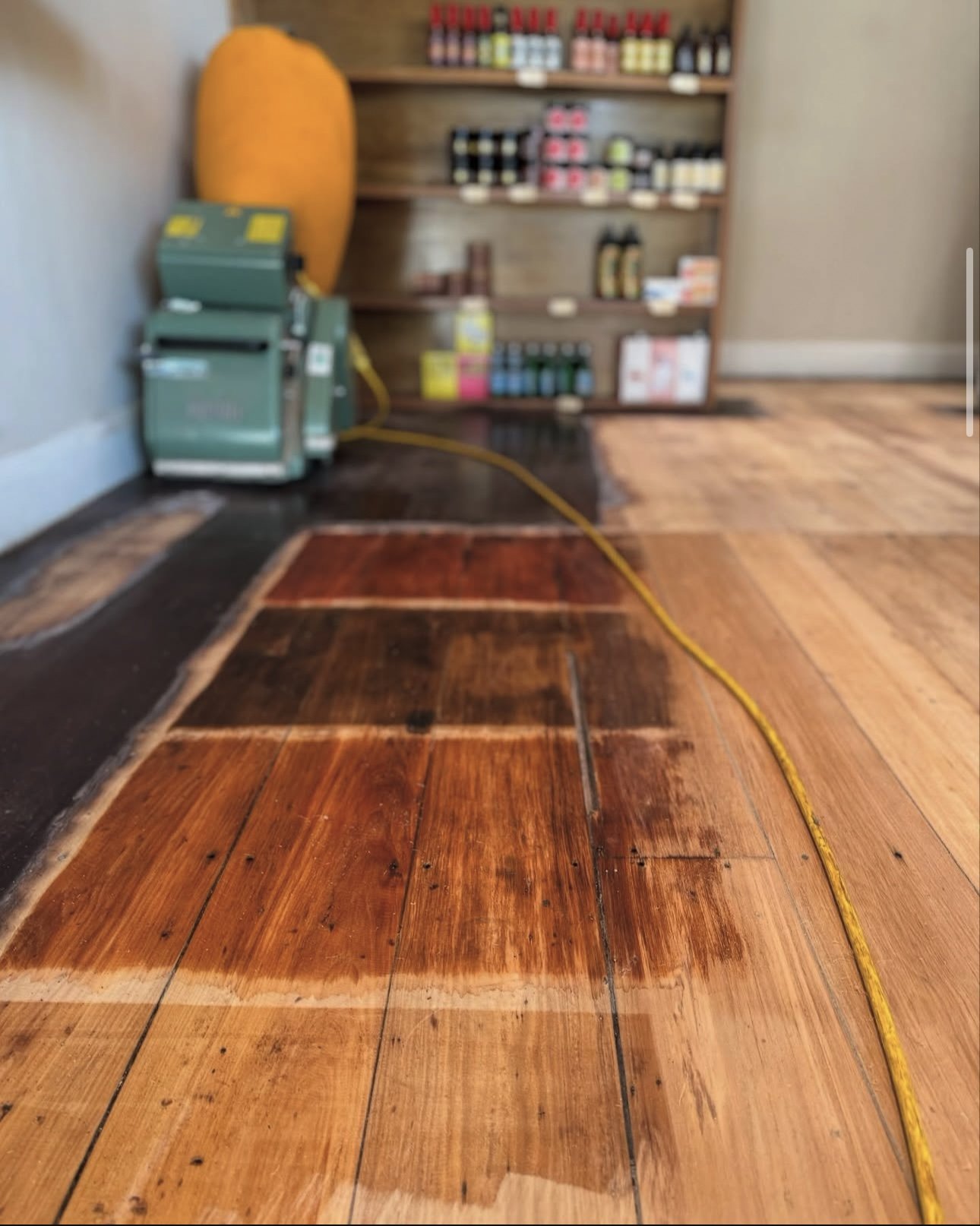 Refinishing hardwood floor with section of partially sanded and stained wood, a yellow extension cord, and a green floor sander machine in a room with a wooden cabinet in the background.