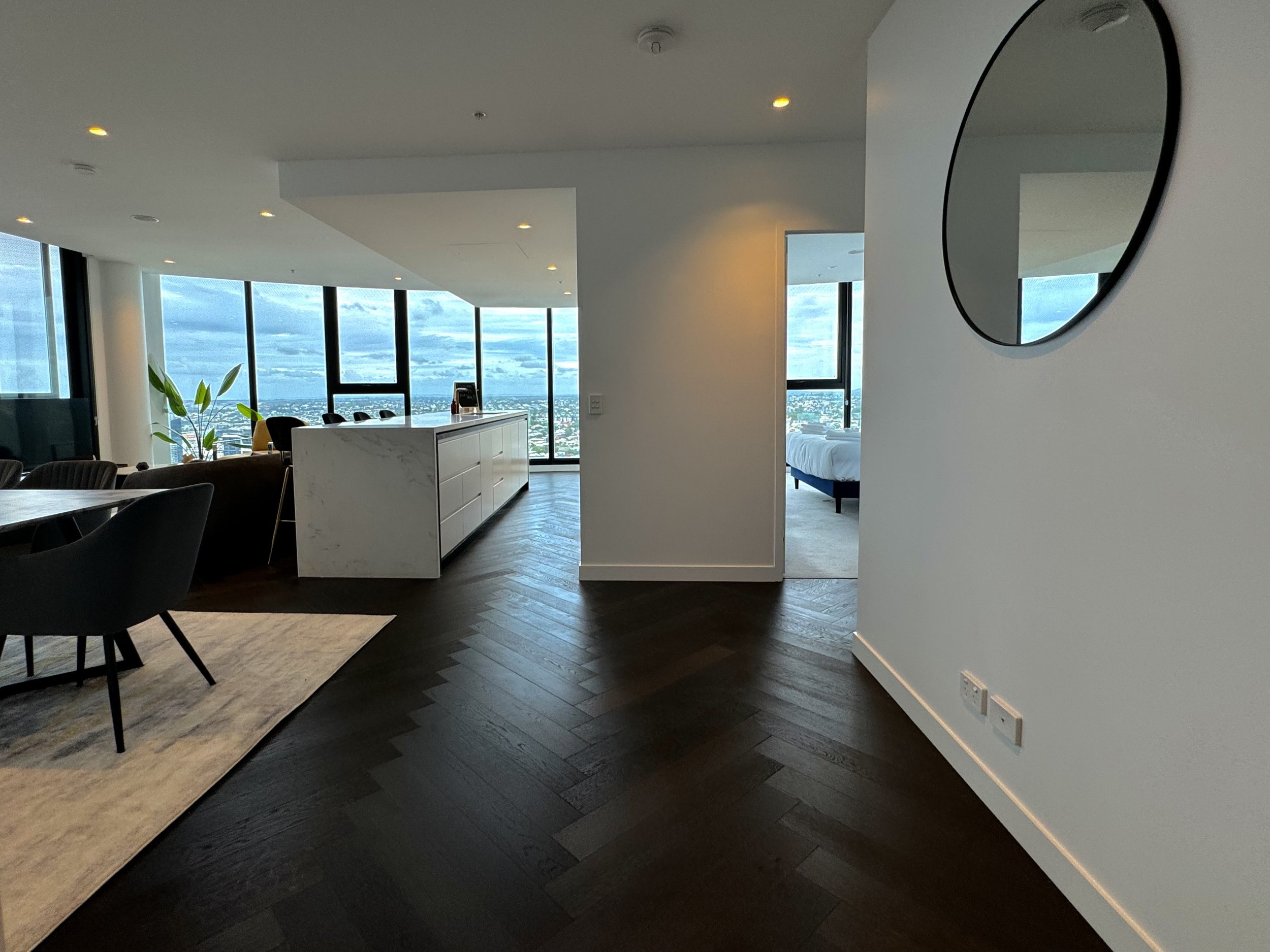 Modern apartment interior with dark hardwood floors, a sitting area with black chairs and a dining table, a white marble-topped island, large windows with a city view, and a bedroom visible through an open doorway.