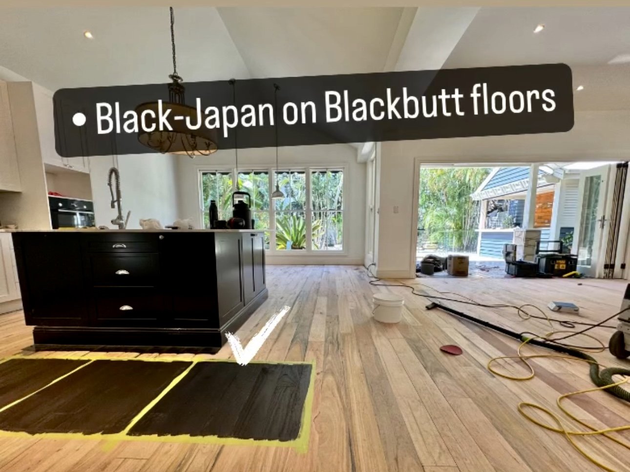 Interior of a house with hardwood floors being refinished with black stain, setup for flooring work surrounded by tools and equipment, large windows and sliding glass door showing garden outside.
