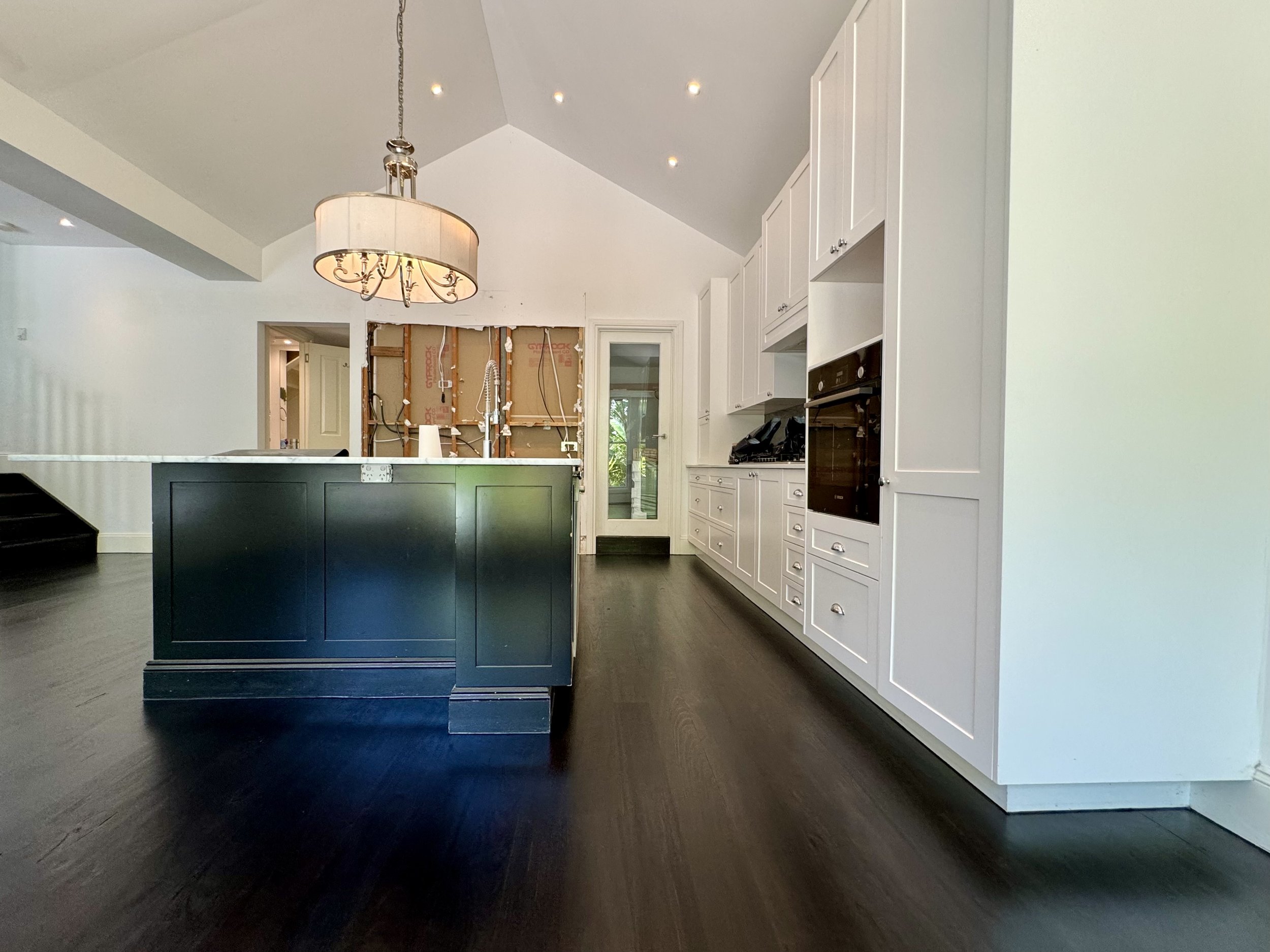 Modern kitchen with white cabinets, black island, dark hardwood flooring, and a chandelier, with an unfinished wall area in the background.