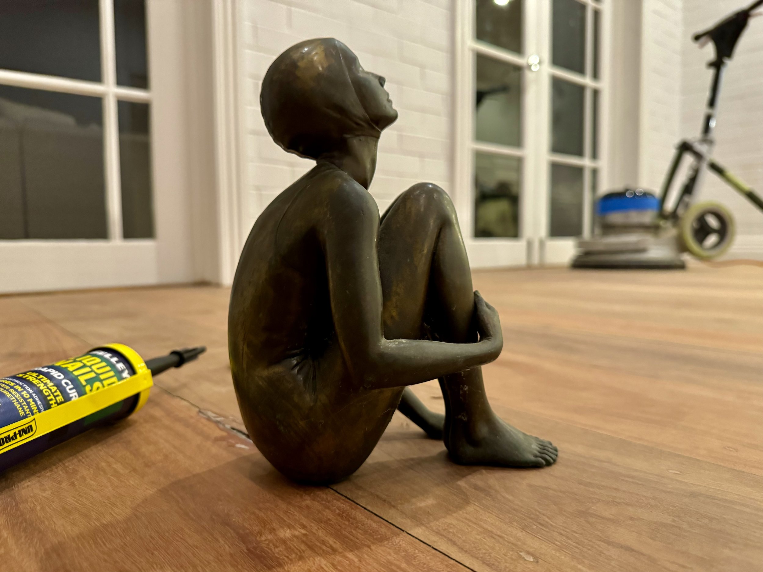 Bronze sculpture of a seated woman with knees drawn to her chest, on a wooden surface with a tube of glue and a skateboard in the background.