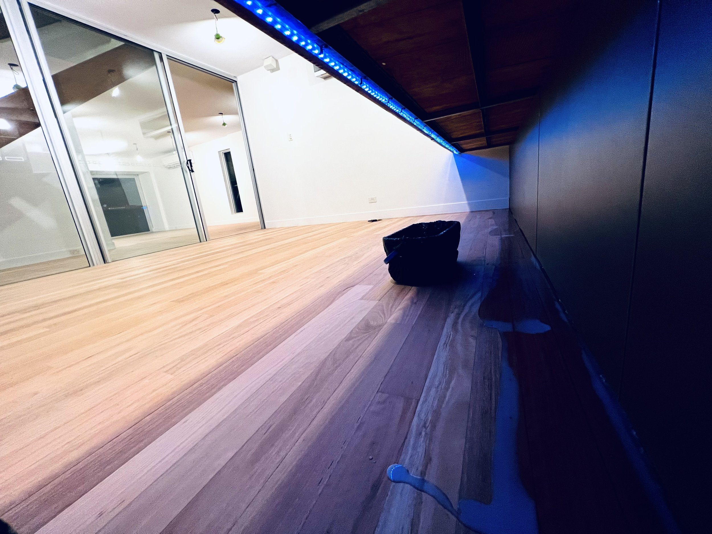 Interior view of a modern room with wooden flooring, white walls, and glass sliding doors reflecting the room, with blue LED strip lighting under a wood-paneled ceiling. A black bag sits on the floor near the wall.