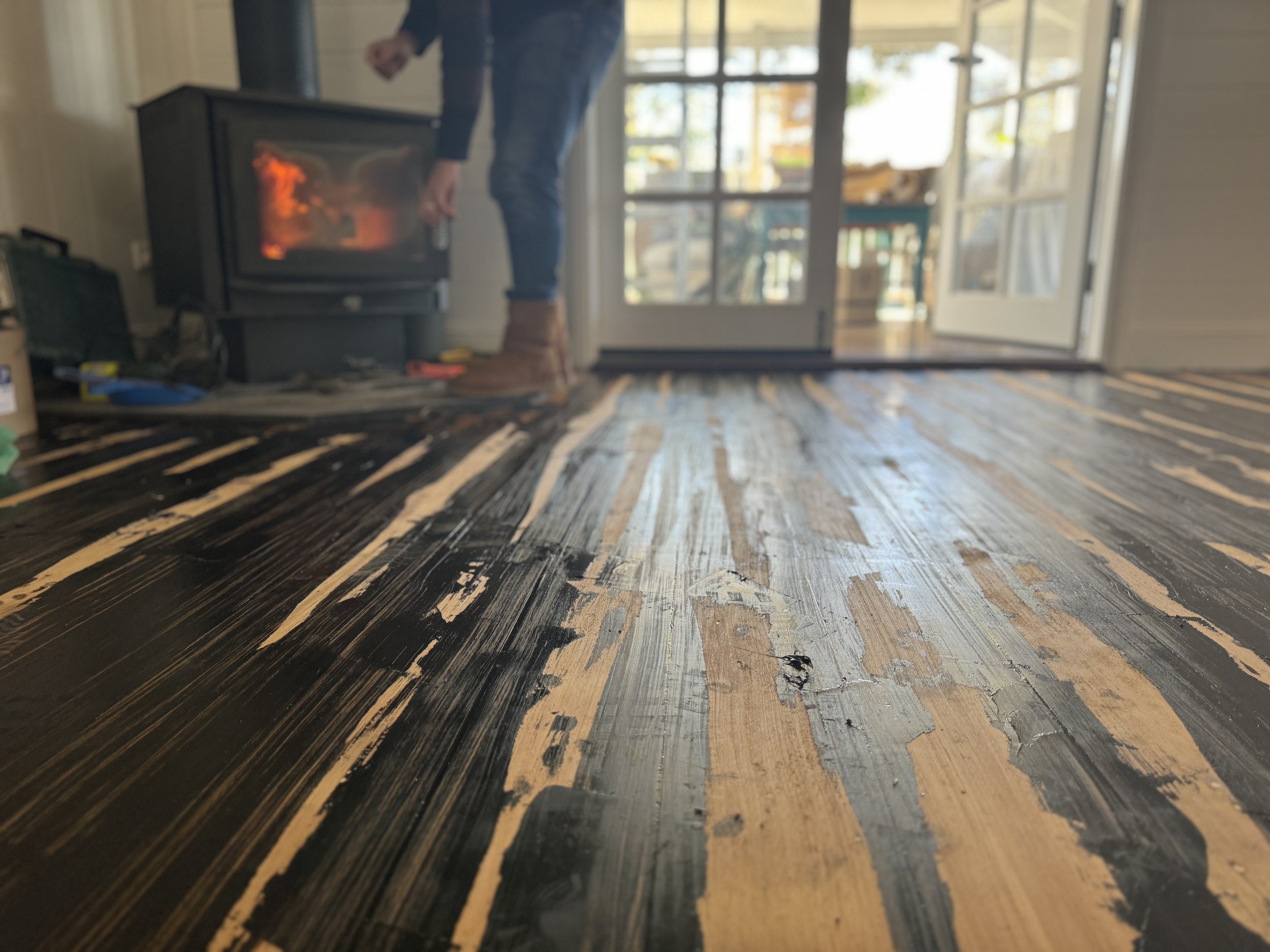 Close-up of a distressed wooden floor with black and natural wood streaks, with a person in the background tending a fireplace or stove through a glass door, and glass doors leading to the outside.