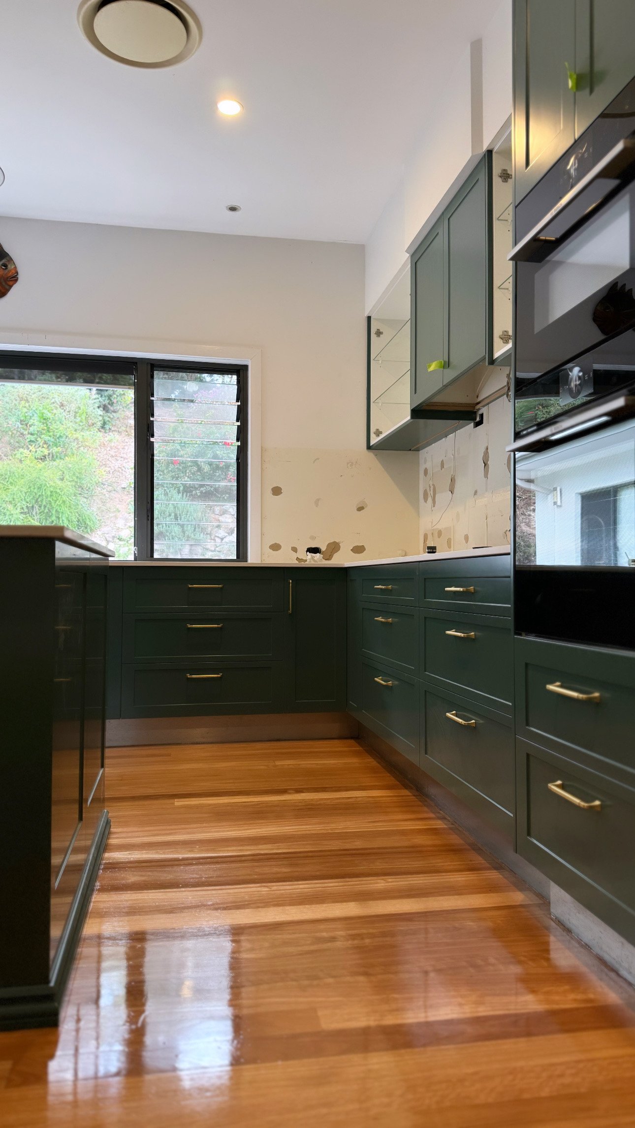 Partially renovated kitchen with green cabinets, hardwood floor, and a window showing trees outside.