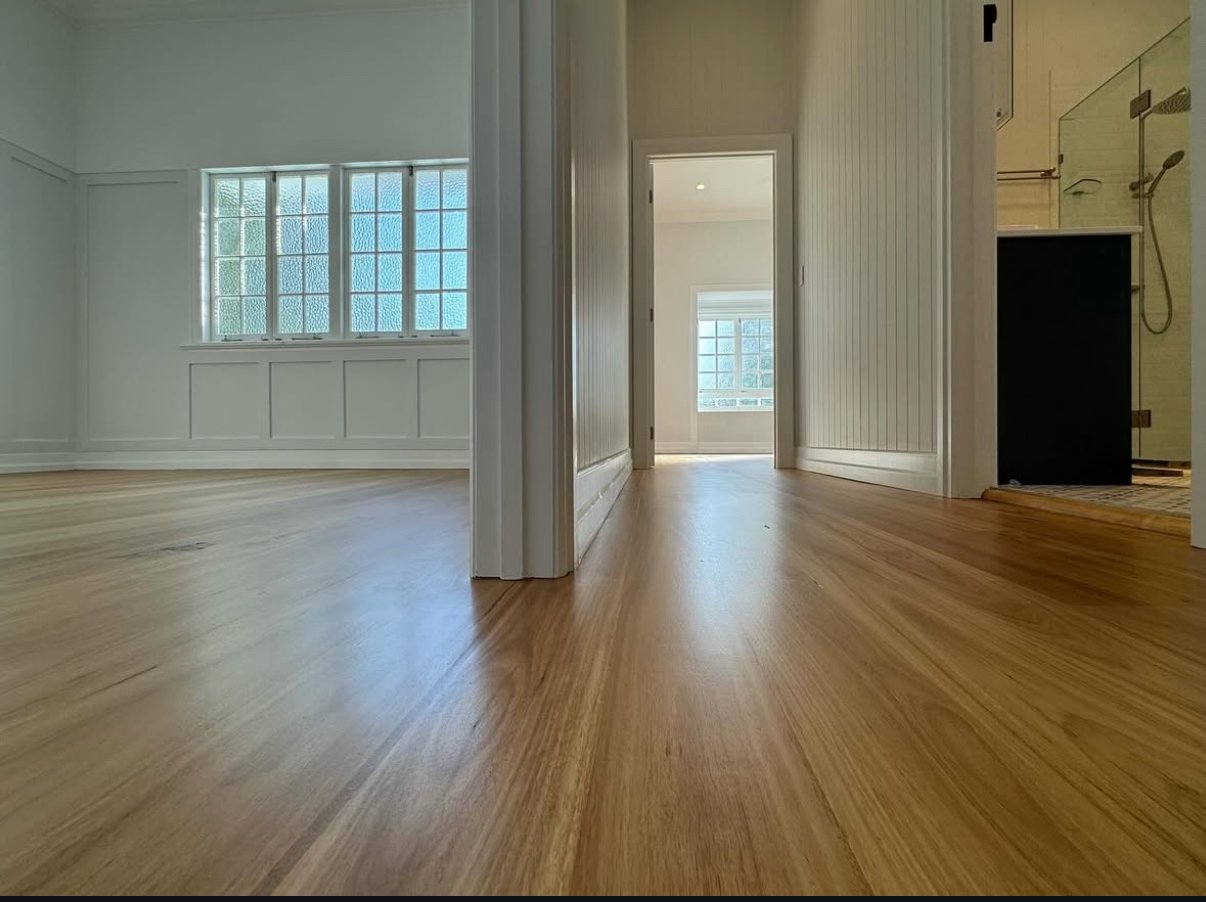 Empty interior rooms of a house with hardwood floors, white walls, large windows, unclear doorways, and a bathroom with a shower.