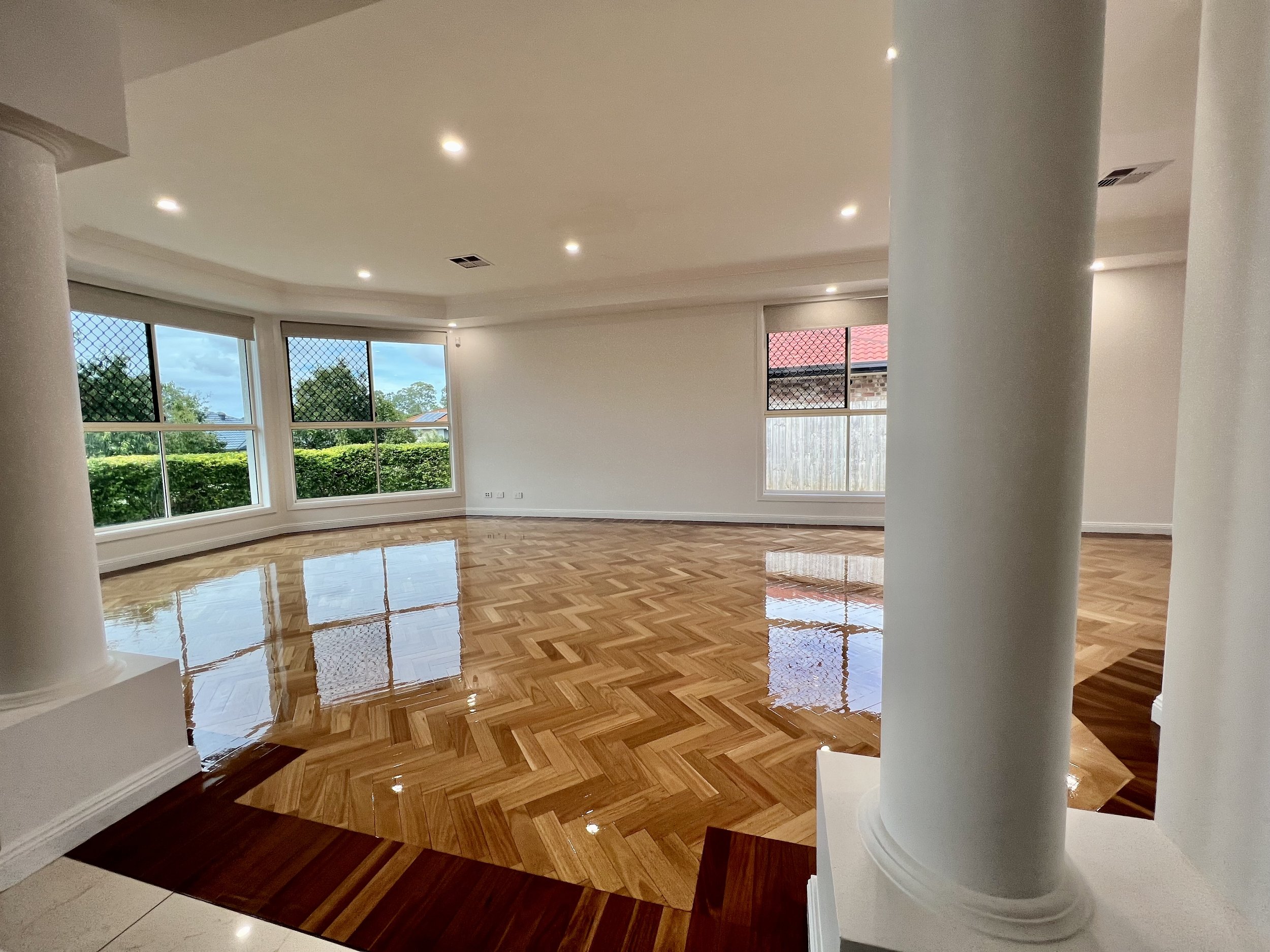 Empty living room with large bay windows, polished wooden floors, and white walls, with two white columns visible in the foreground.
