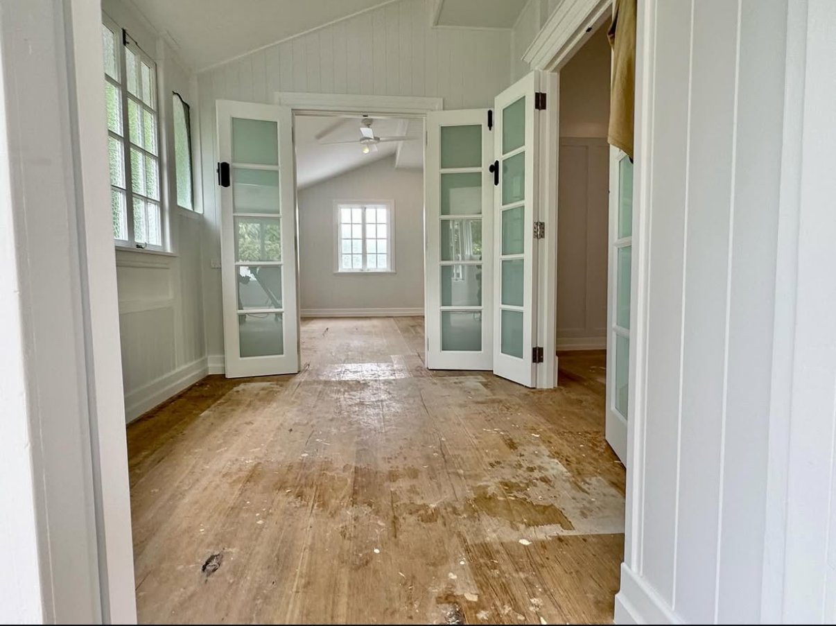 Empty room with unfinished wooden floor, white walls, and multiple windows. French doors lead to another room with a ceiling fan.