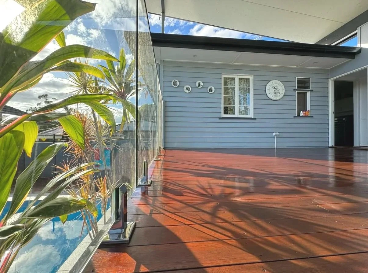 Re-coated deck seen through a glass pool fence, palm shadows falling across the boards