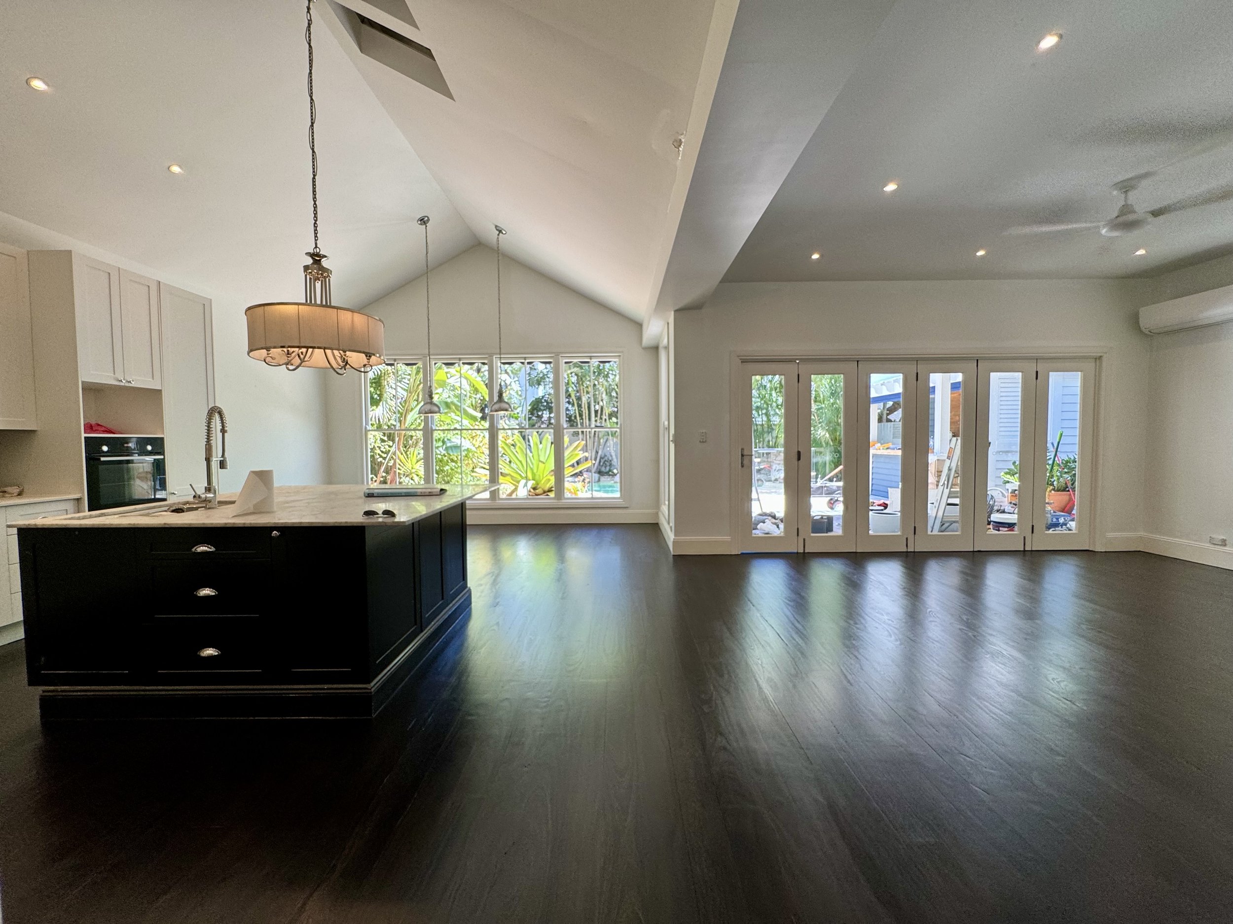 Empty living room with dark hardwood floors, white walls, large glass doors to the outside, and a kitchen island with a beige countertop and black base.