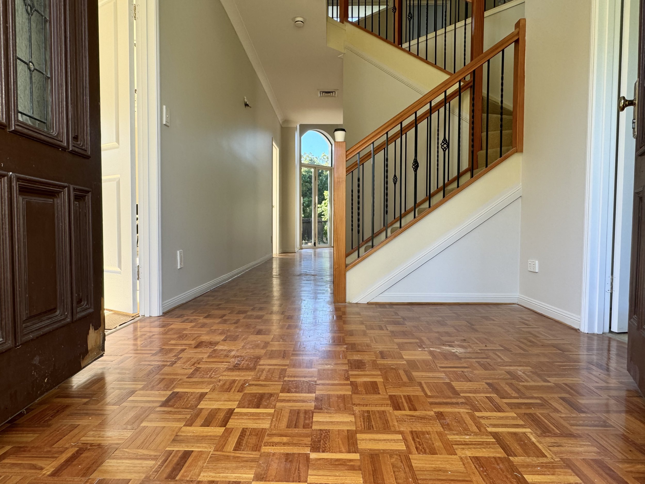 Interior view of a house entrance with hardwood floors, an open front door, stairs leading upstairs with wooden handrails and black balusters, and a hallway with a door at the end showing greenery outside.