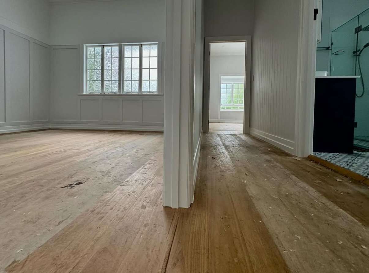 Interior of a house under renovation, showing empty rooms with unfinished wood floors, white walls, and windows with textured glass. There is an open doorway leading to a bathroom with a glass shower enclosure.