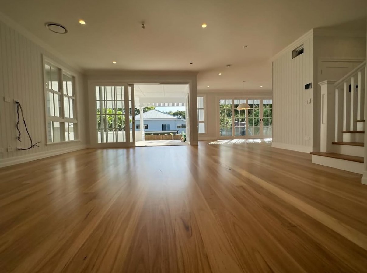 Empty living room with hardwood floors, large windows, and a sliding glass door leading outside, with sunlight streaming in.