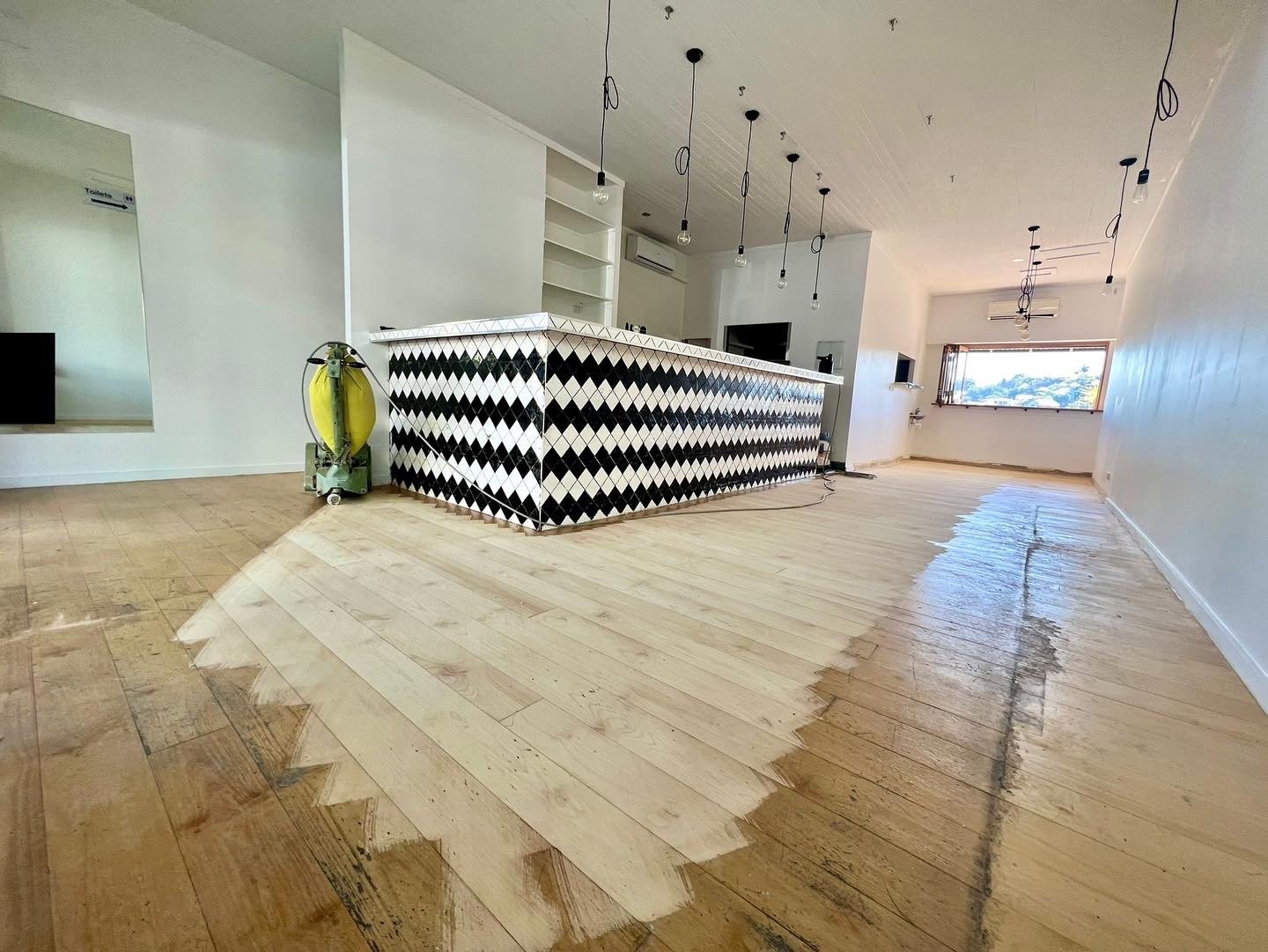 Interior of a room under renovation with a partially painted hardwood floor, a patterned bar front, and hanging light fixtures.