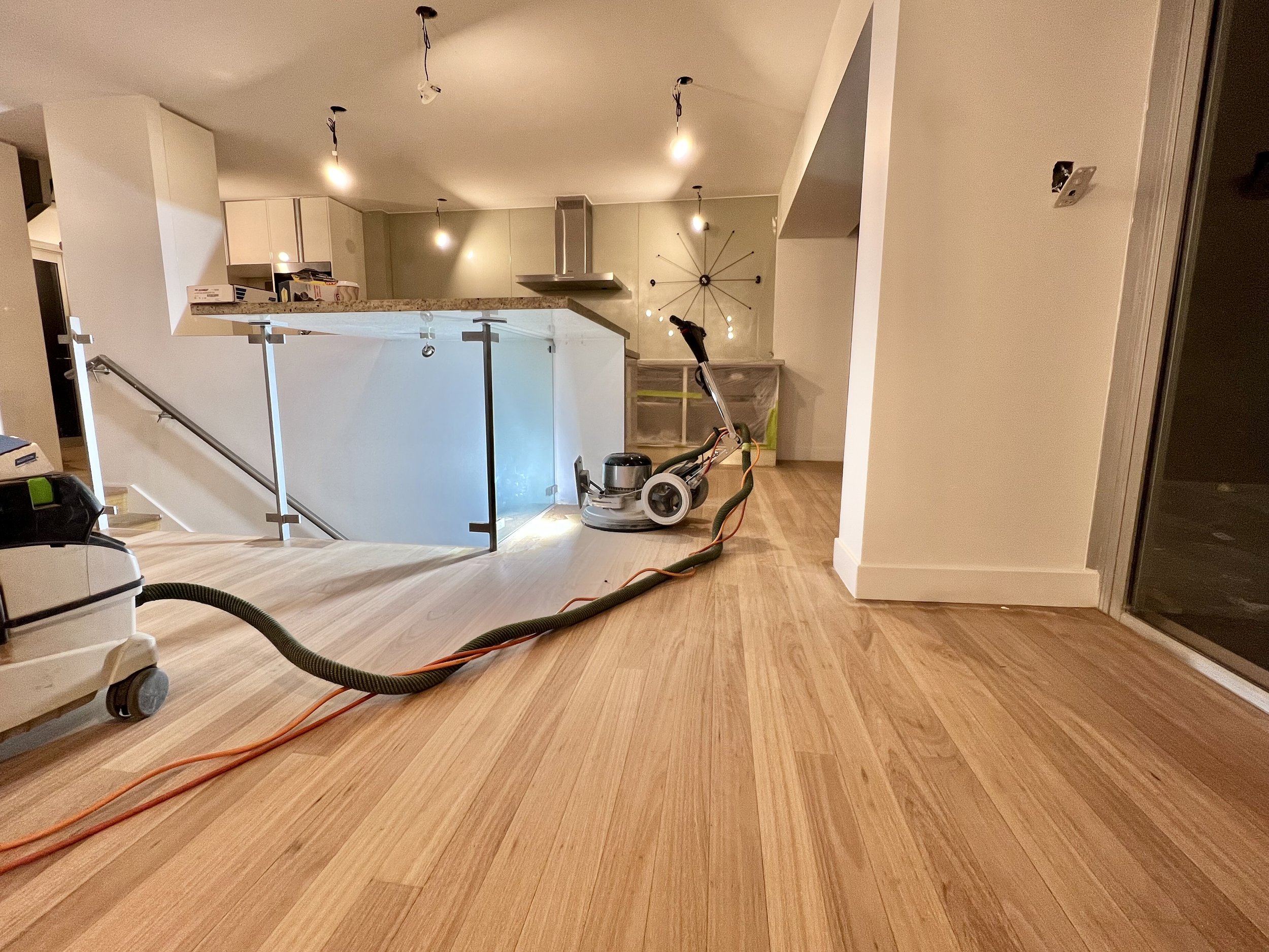 Living room with hardwood floors, a staircase with glass railing, and a partially visible kitchen in the background. Construction equipment and a floor sander are present, indicating ongoing renovation.