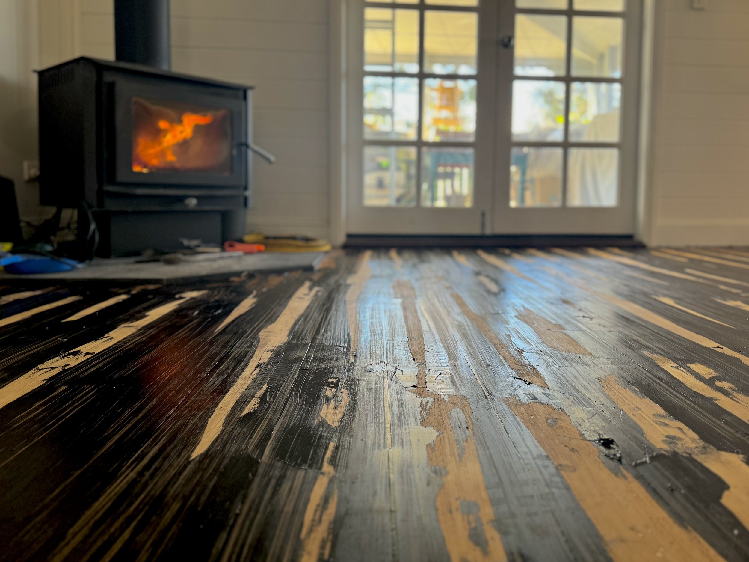 Close-up of a wooden floor with black and natural wood streaks, showing a glossy finish and some minor scratches. In the background, there are glass double doors leading outside and a black wood stove with a fire inside.