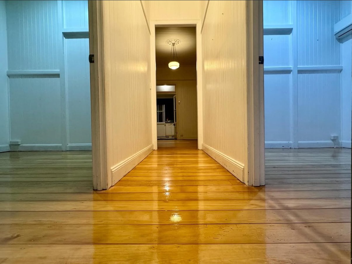 Image of a hallway with wooden floors and white paneled walls, leading to a room at the end with a ceiling light, seen from a low angle.