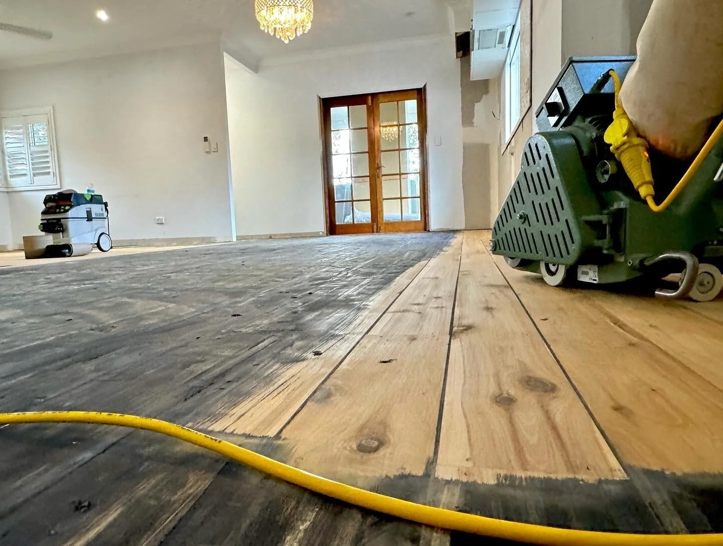 Interior of a room under renovation showing unfinished hardwood floor, window shutters, a door with glass panes, and construction equipment including a green floor sander and a vacuum, with some flooring partially sanded.