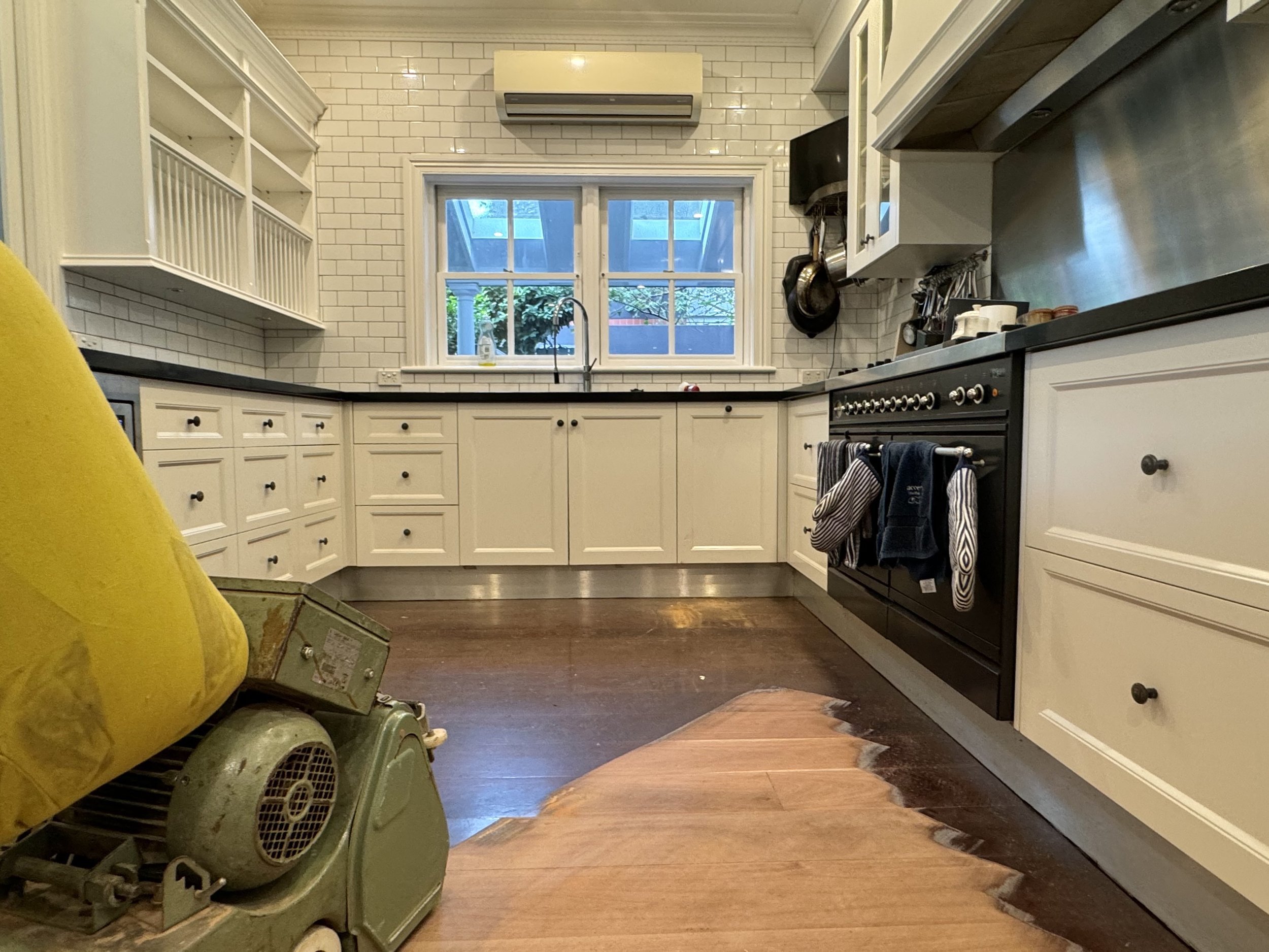 Kitchen with white cabinets, black countertops, a stove with oven and hanging towels, a window above the sink showing an outdoor patio, and a partially removed wooden floor section revealing the subfloor.
