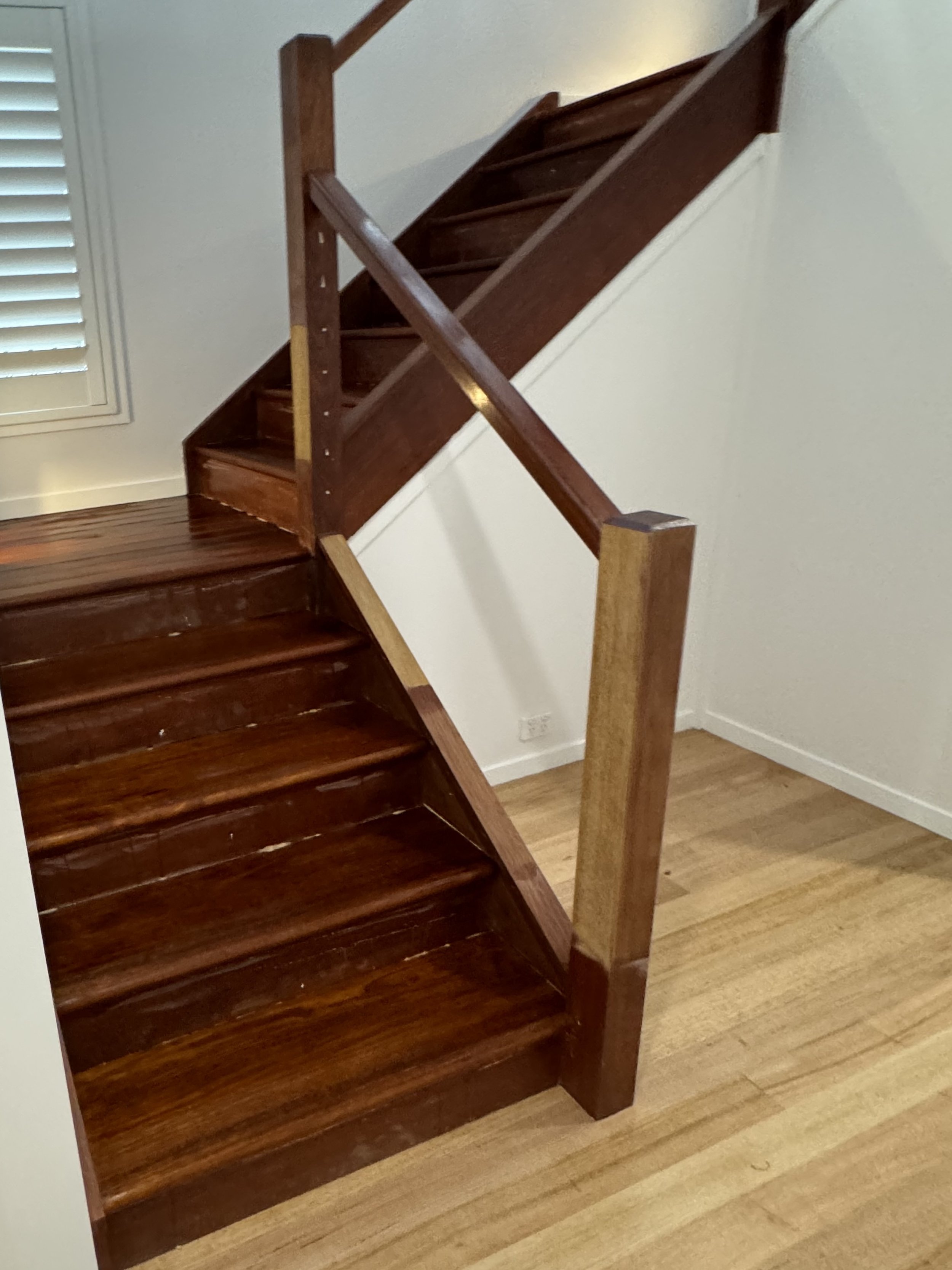 Wooden staircase with dark stained steps and a light wood handrail, shown from the bottom, near a window with white shutters.