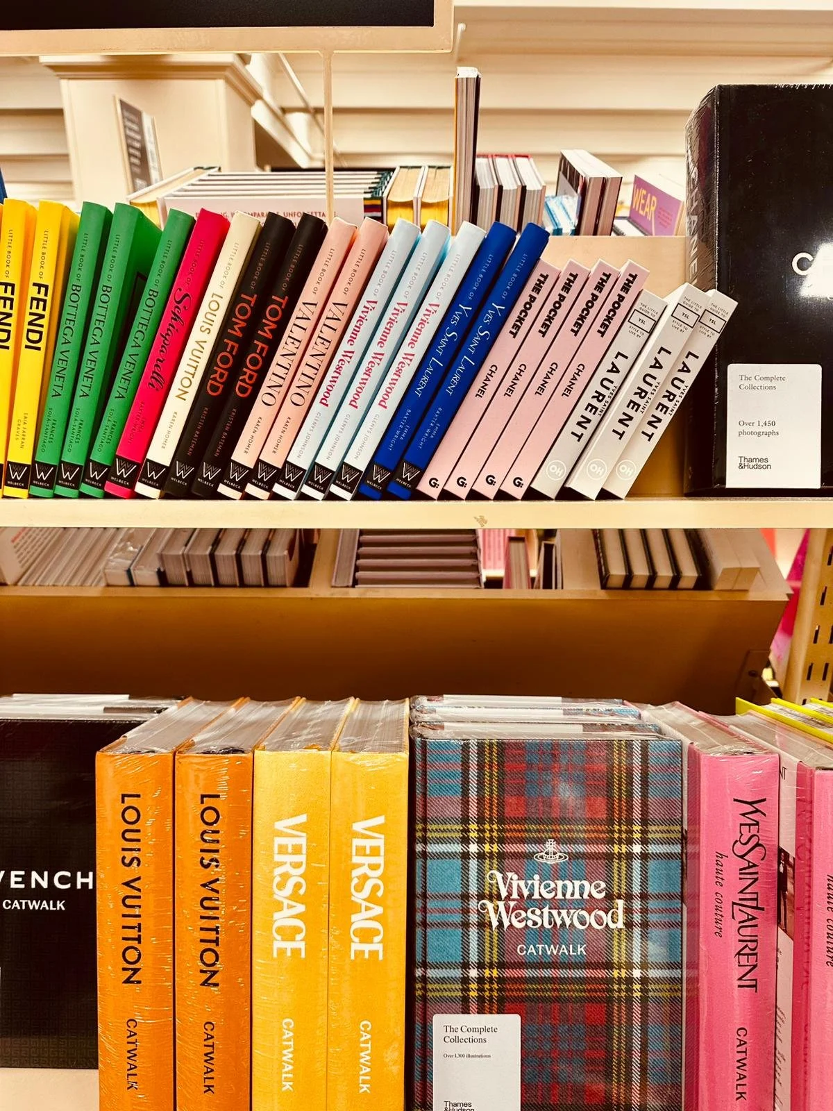 Bookshelves filled with various books, including titles by Louis Vuitton, Tom Ford, Vivienne Westwood, Yves Saint Laurent, and Chanel, arranged in a display at a bookstore or shop.