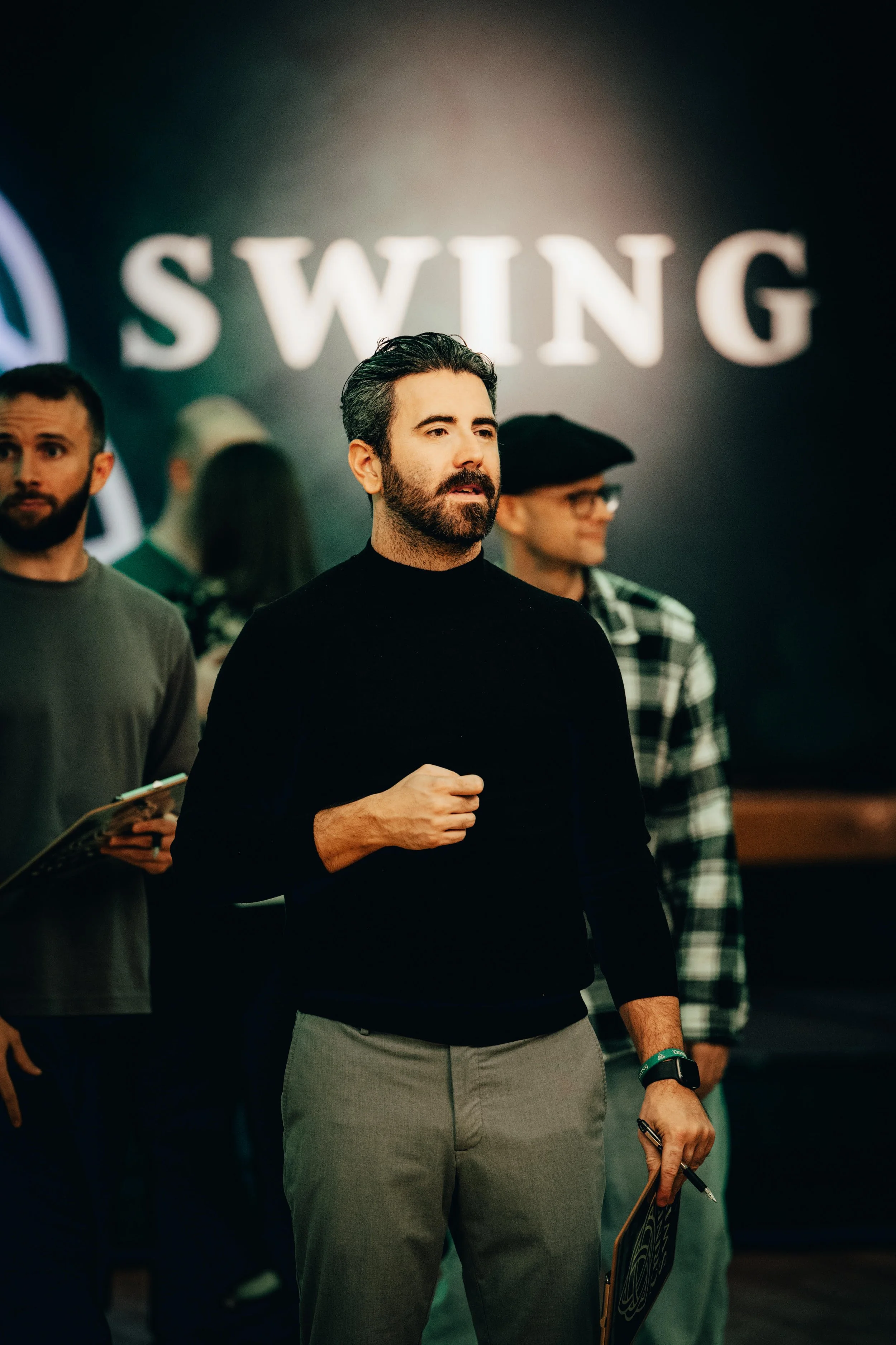 Hombre de cabello oscuro y barba, vistiendo un suéter negro y pantalones grises, sosteniendo un bolígrafo y una libreta en una sala con personas y un letrero grande que dice 'SWING' al fondo.