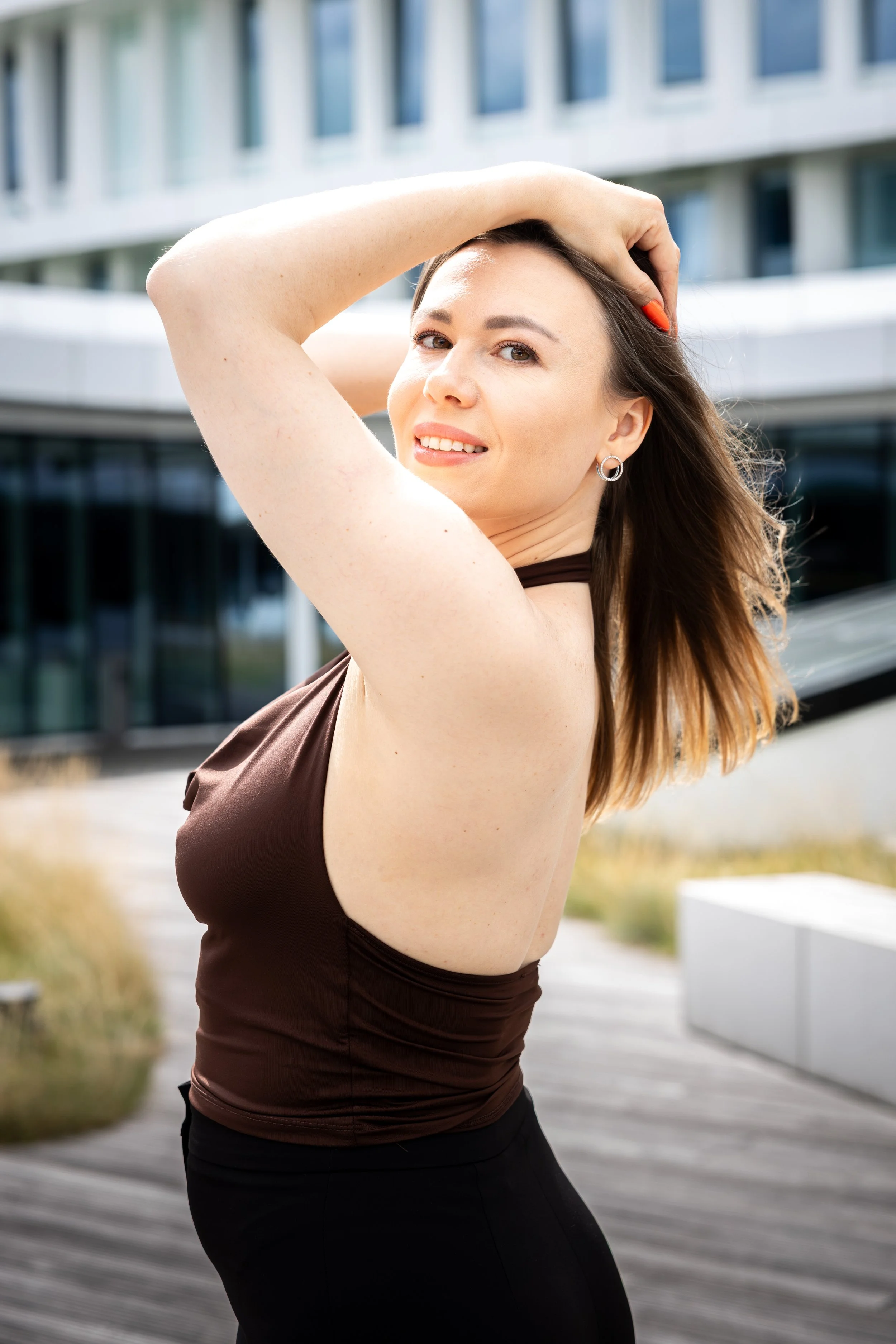 Mujer sonriente con cabello largo y camisa marrón, posando con una mano en la cabeza frente a un edificio moderno con ventanas de vidrio.