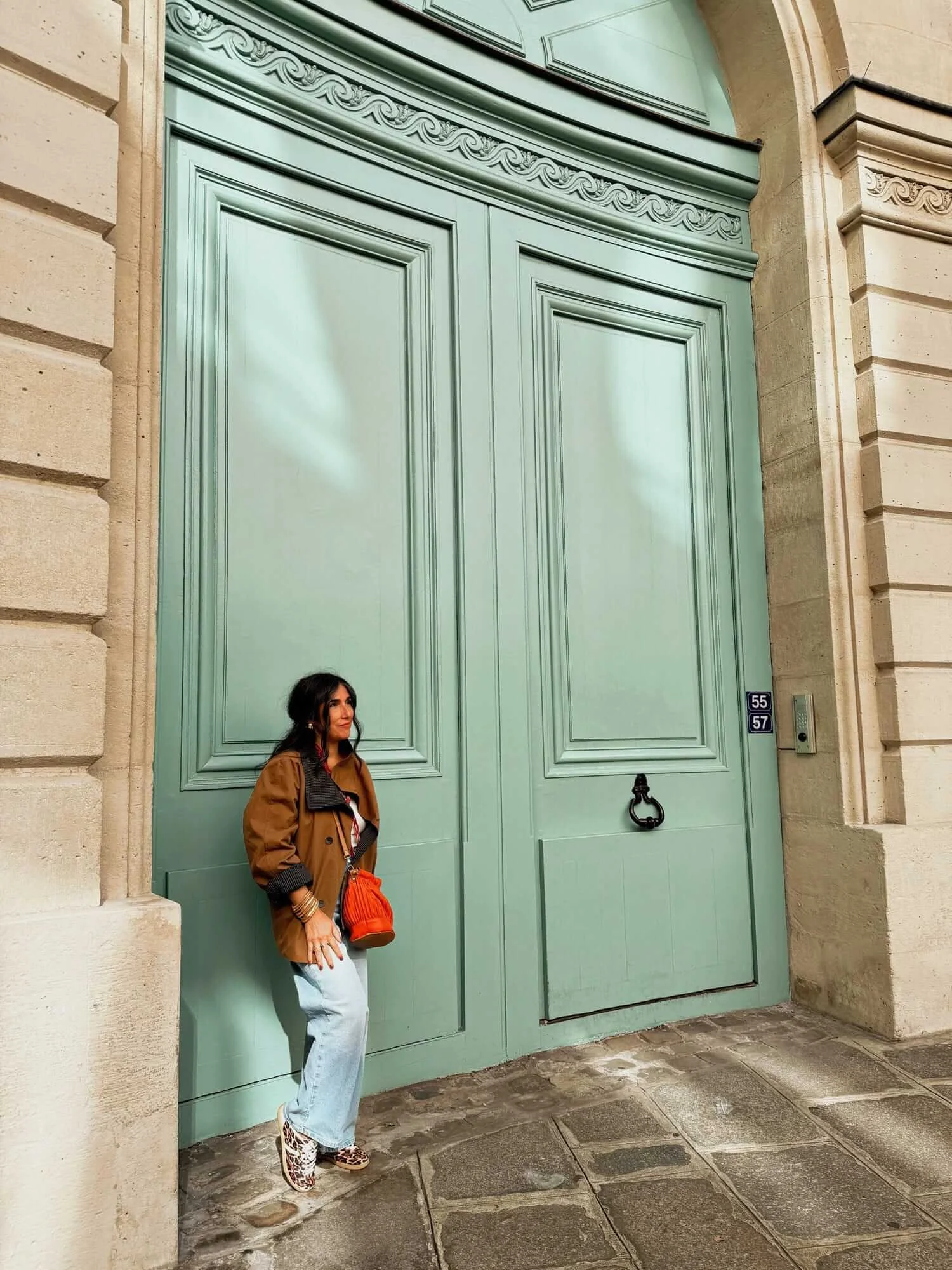 Elizabeth Quintal web designer in jeans, brown jacket, leaning against large teal doors and smiling