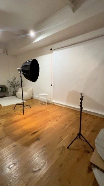 Ein Foto eines leeren Fotostudios mit professionellem Licht- und Stativausrüstung auf Holzboden, weißen Wänden und einem großen weißen Hintergrund. Es gibt auch Möbel und Pflanzen im Hintergrund.