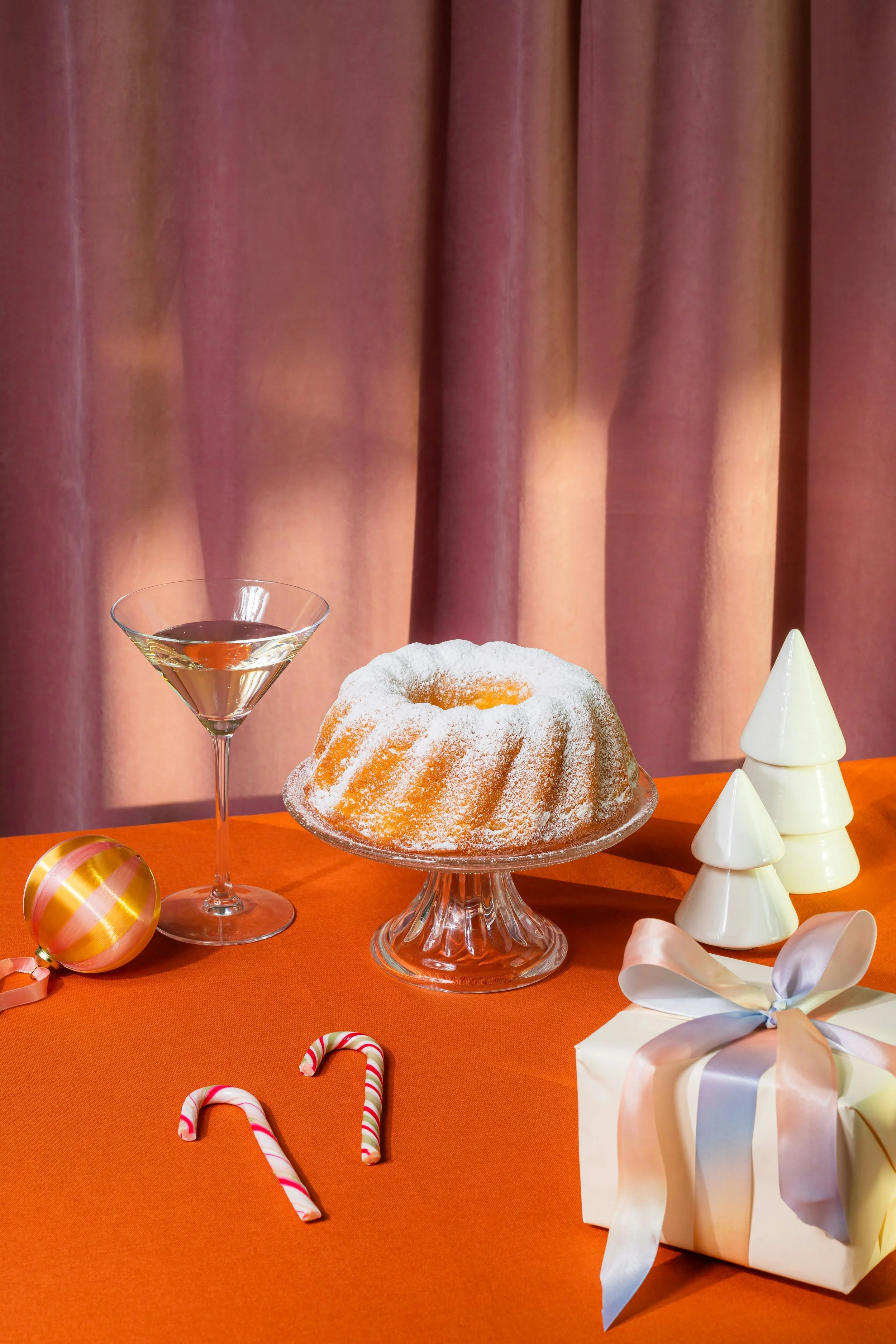 Festliche Weihnachtsdekoration mit Kuchen, Geschenk, Weihnachtskugeln, Zuckerstangen, Schneeflocken und Tannenzapfen auf orangefarbenem Tischtuch vor rosa Vorhängen.