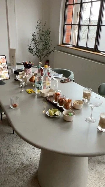 Ein Esstisch mit verschiedenen Getränken, Snacks, Gläsern und Tellern in einem hellen Raum mit Fenster und Pflanze.