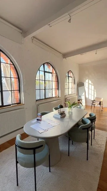 Ein heller Konferenzraum mit großen, rundbogenförmigen Fenstern, einem ovalen Tisch mit Stühlen, Unterlagen und Pflanzen, und einem großen Spiegel an der Wand.