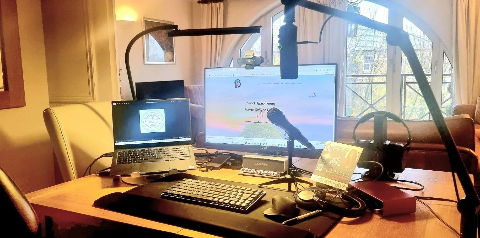 A workspace with a computer monitor, notebook, and microphone set up on a wooden table. The monitor displays a website titled "Synct Hypnotherapy." There is a laptop, a small Bose speaker, headphone, and various electronic devices on the table. Behind, a large window with curtains and a sofa can be seen.
