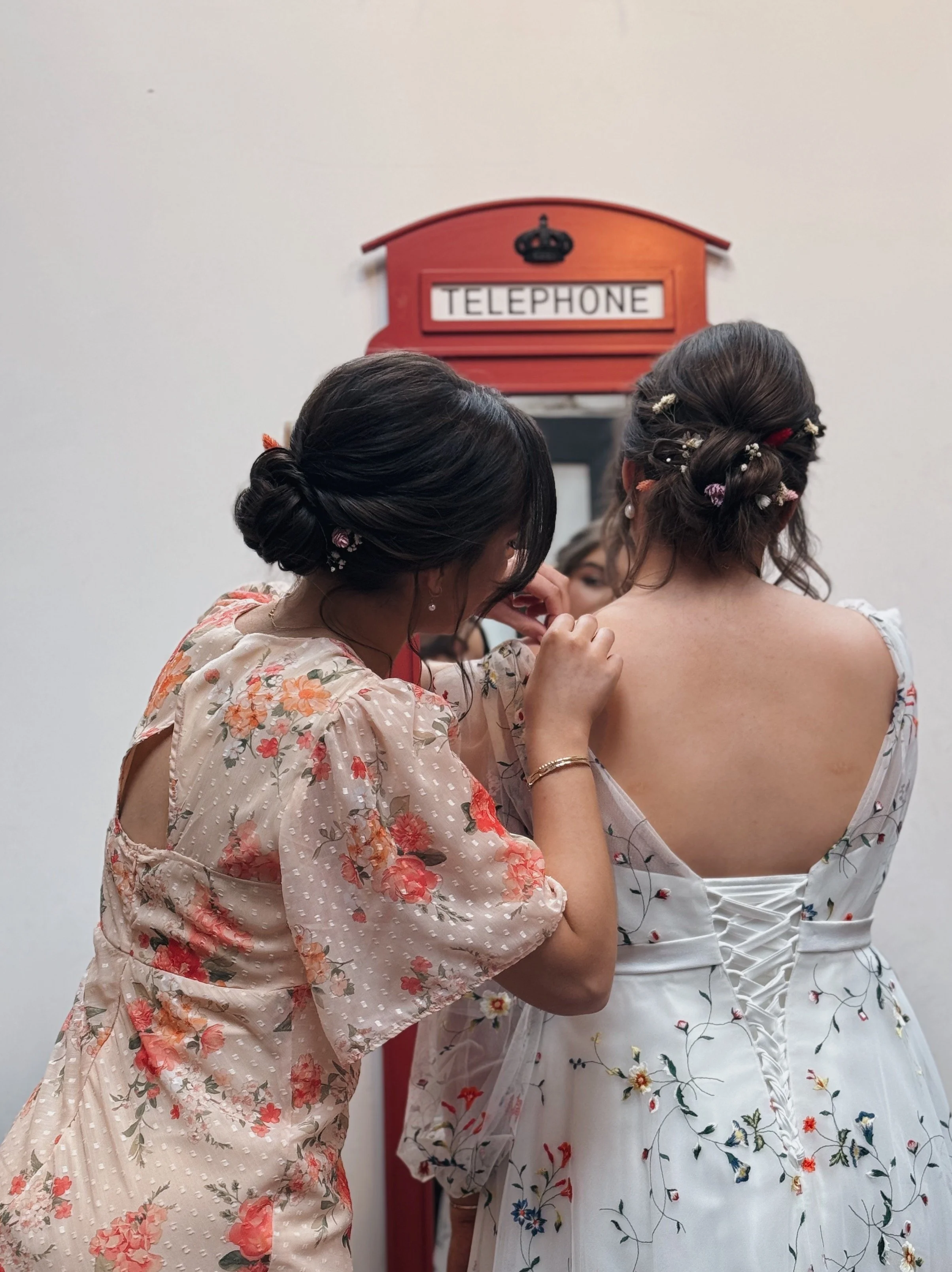 A woman in a floral dress helps another woman in a floral wedding gown with her dress. The wedding gown has a lace-up back. They are in a room with a red vintage telephone booth mirror on the wall, Comrie Croft, Perthshire, Scotland.