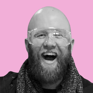 A smiling man with a beard wearing glasses and a scarf, against a pink background.