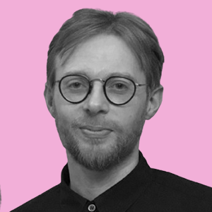 A man with glasses and a beard, wearing a black shirt, standing against a pink background.