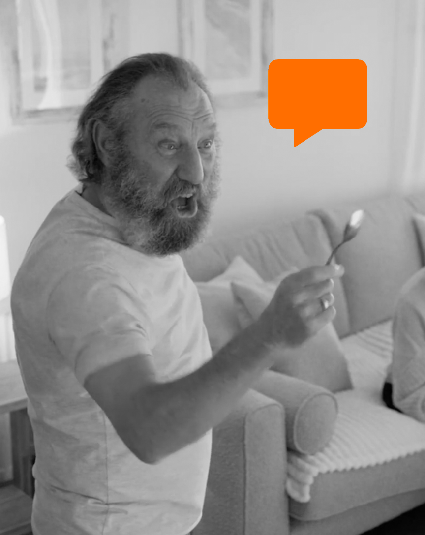 A bearded man with gray hair, wearing a light-colored t-shirt, sitting on a couch, holding a spoon, and speaking animatedly. An orange speech bubble appears above his head.