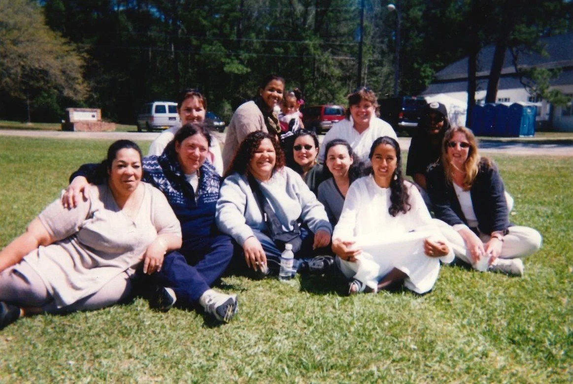 A group of eleven women and children sitting and standing on grass outdoors with trees, parked cars, and portable toilets in the background.