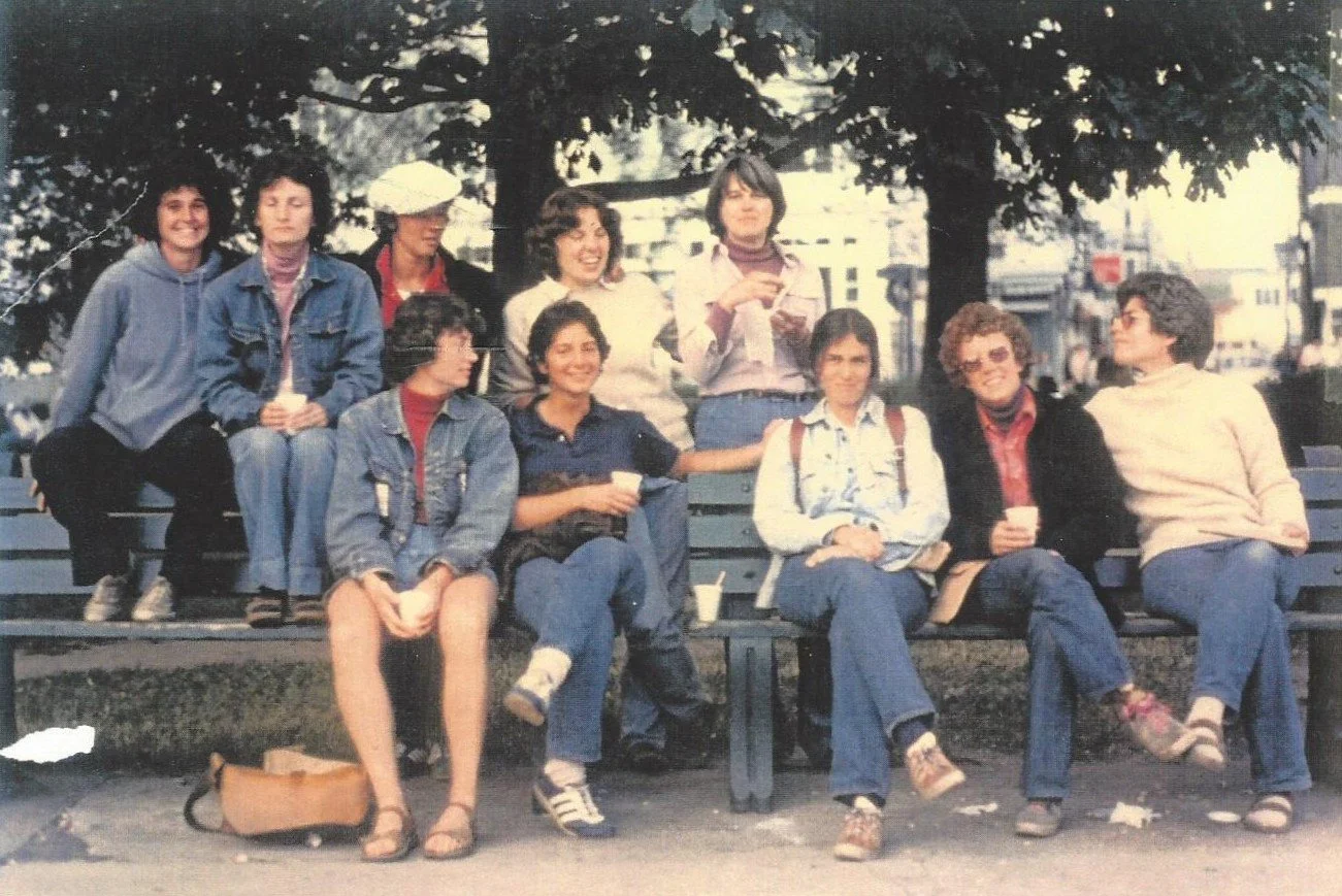 A group of ten women sitting and standing on a park bench with trees and buildings in the background.
