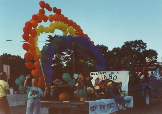 Group of people on a decorated parade float with colorful balloons in red, orange, yellow, green, and blue, and a sign during a daytime parade.
