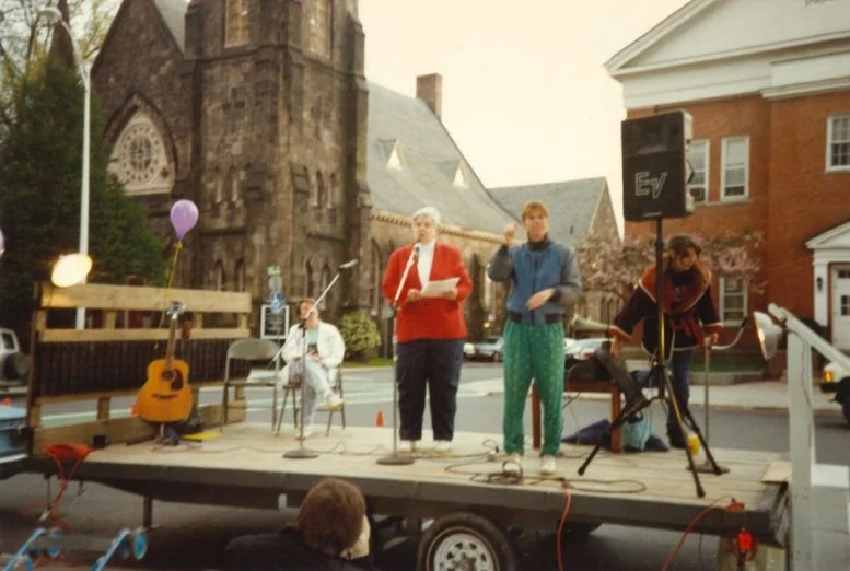 People performing on a small outdoor stage in front of a church with a boy in a red jacket, a girl in a sweater, and a person seated with a guitar. There are microphones and speakers set up, and a person in the audience taking photos.