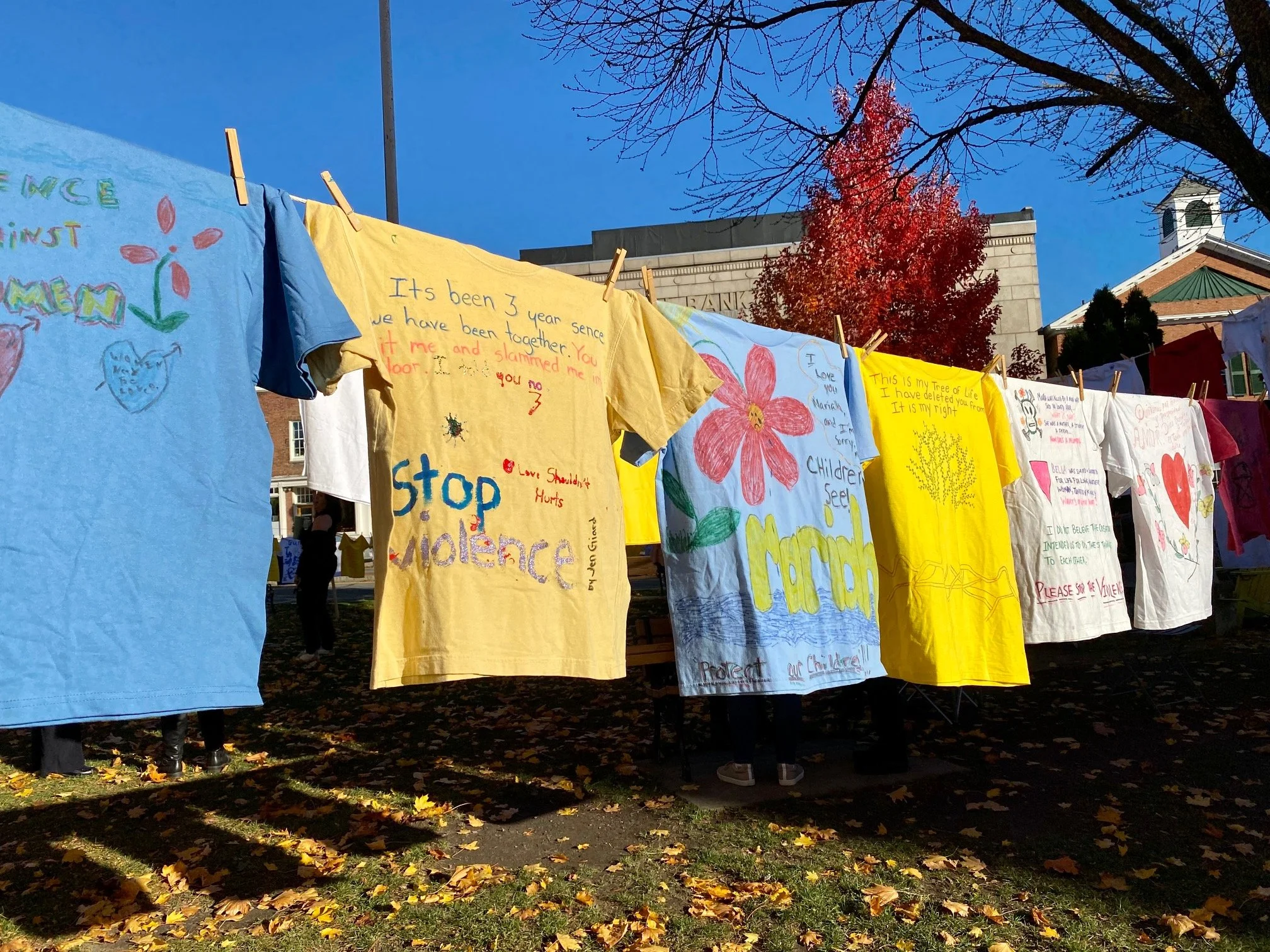 Handmade shirts hanging on a clothesline with messages and drawings, protesting against violence, violence, and promoting kindness, under blue sky and fall trees.