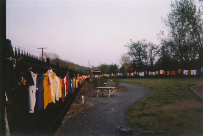 A long line of colorful clothes hanging on a fence outdoors, with a gravel path, picnic table, and trees in the background.