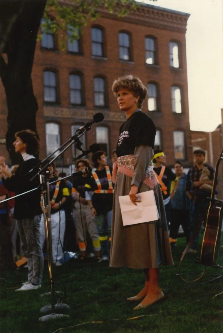 A woman standing at a microphone on a grassy area during a public gathering or protest, holding a piece of paper, with a group of people and buildings in the background.