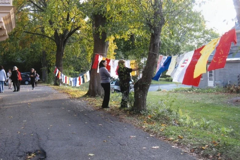 People hanging colorful flags on a line strung between trees in a suburban neighborhood with houses and a street in the background.