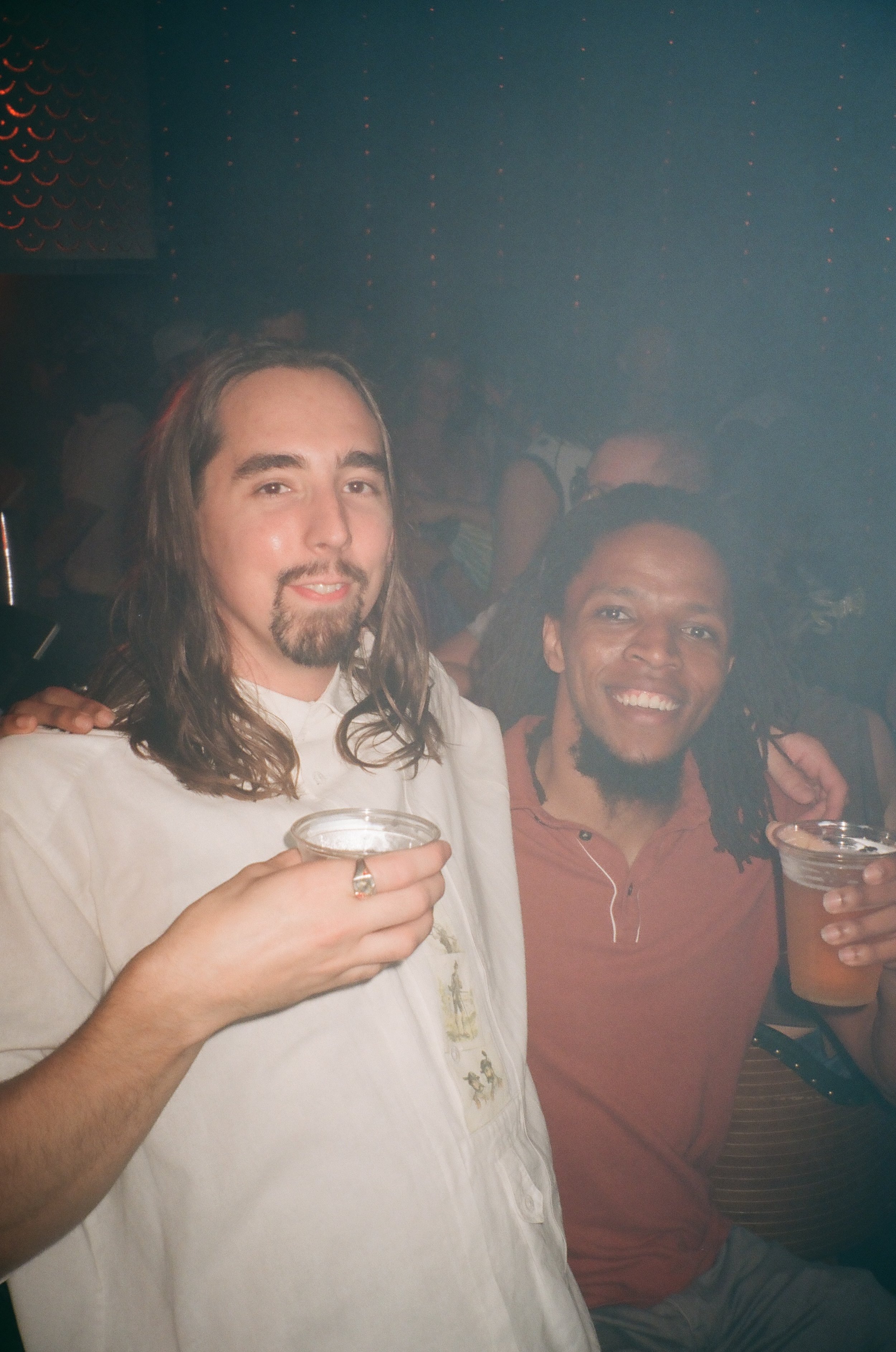 Two men at a party, smiling and holding drinks, with a dark, festive background.
